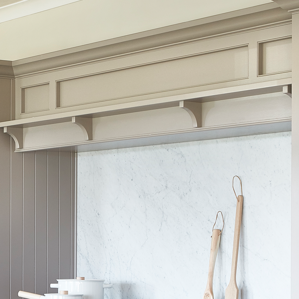 A close-up of a beige kitchen shelf with decorative paneling above a white marble backsplash. Two wooden utensils hang on hooks, and white cookware sits on the countertop below.