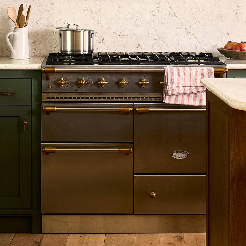 A modern kitchen featuring a stainless steel oven with brass accents, set between dark wood cabinets. A black teapot is on the marble countertop above the oven. Various jars are on the counter in the background.