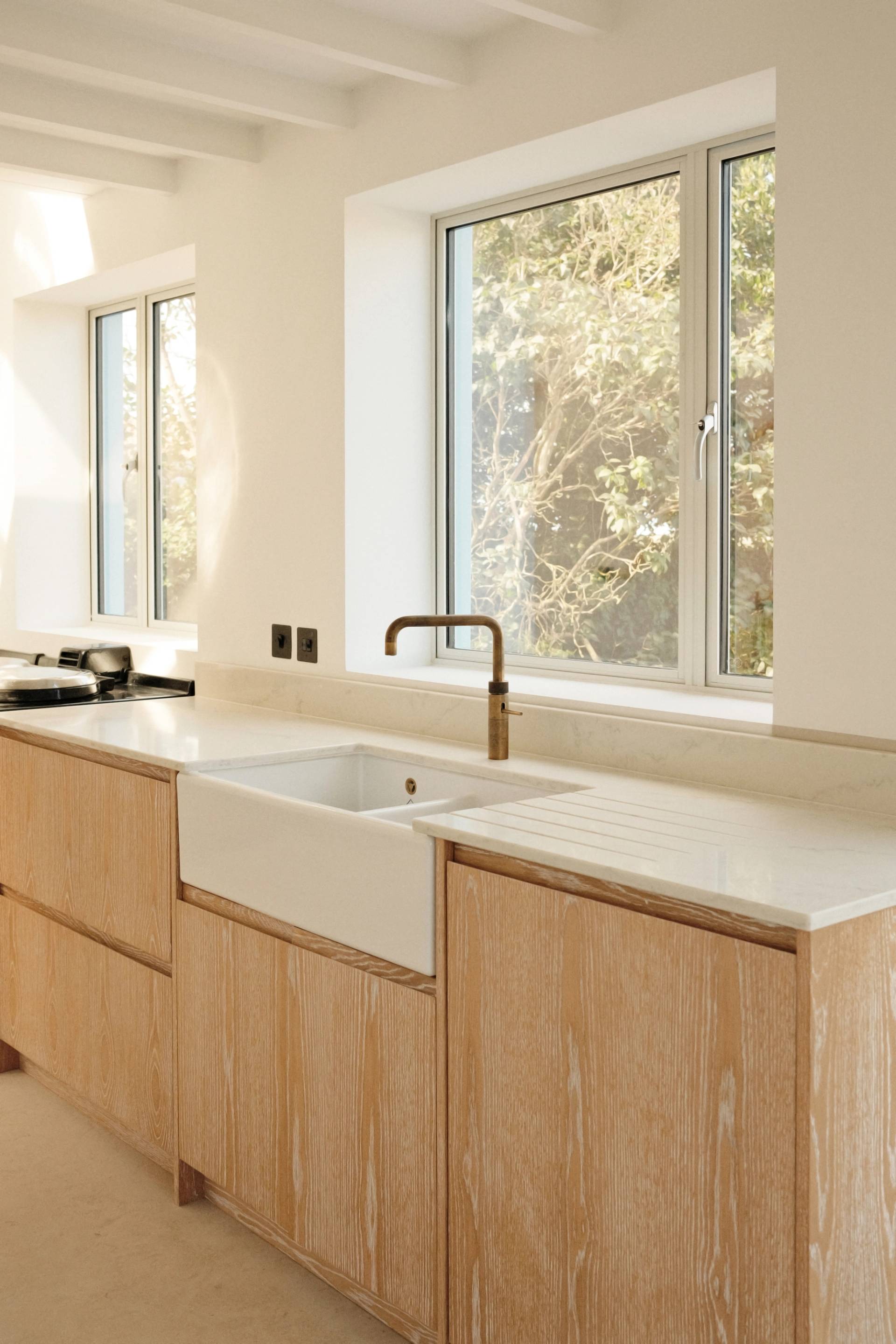 A bright, minimalist kitchen with light wood cabinets, a white farmhouse sink, a brass faucet, and large windows allowing natural light in. The countertop is white, and the walls are painted a soft cream.