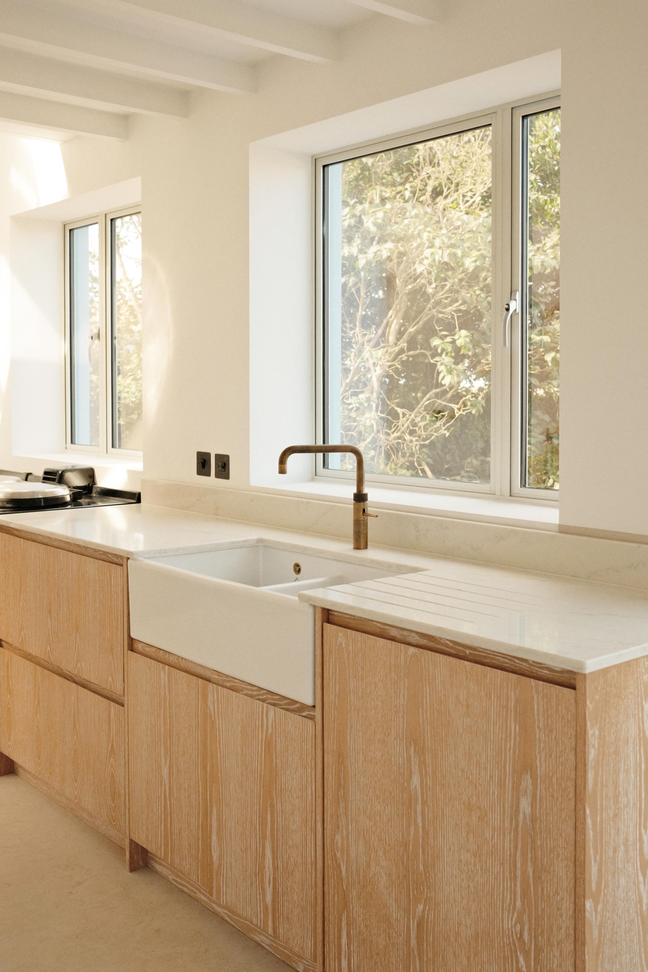 A bright, minimalist kitchen with light wood cabinets, a white farmhouse sink, a brass faucet, and large windows allowing natural light in. The countertop is white, and the walls are painted a soft cream.