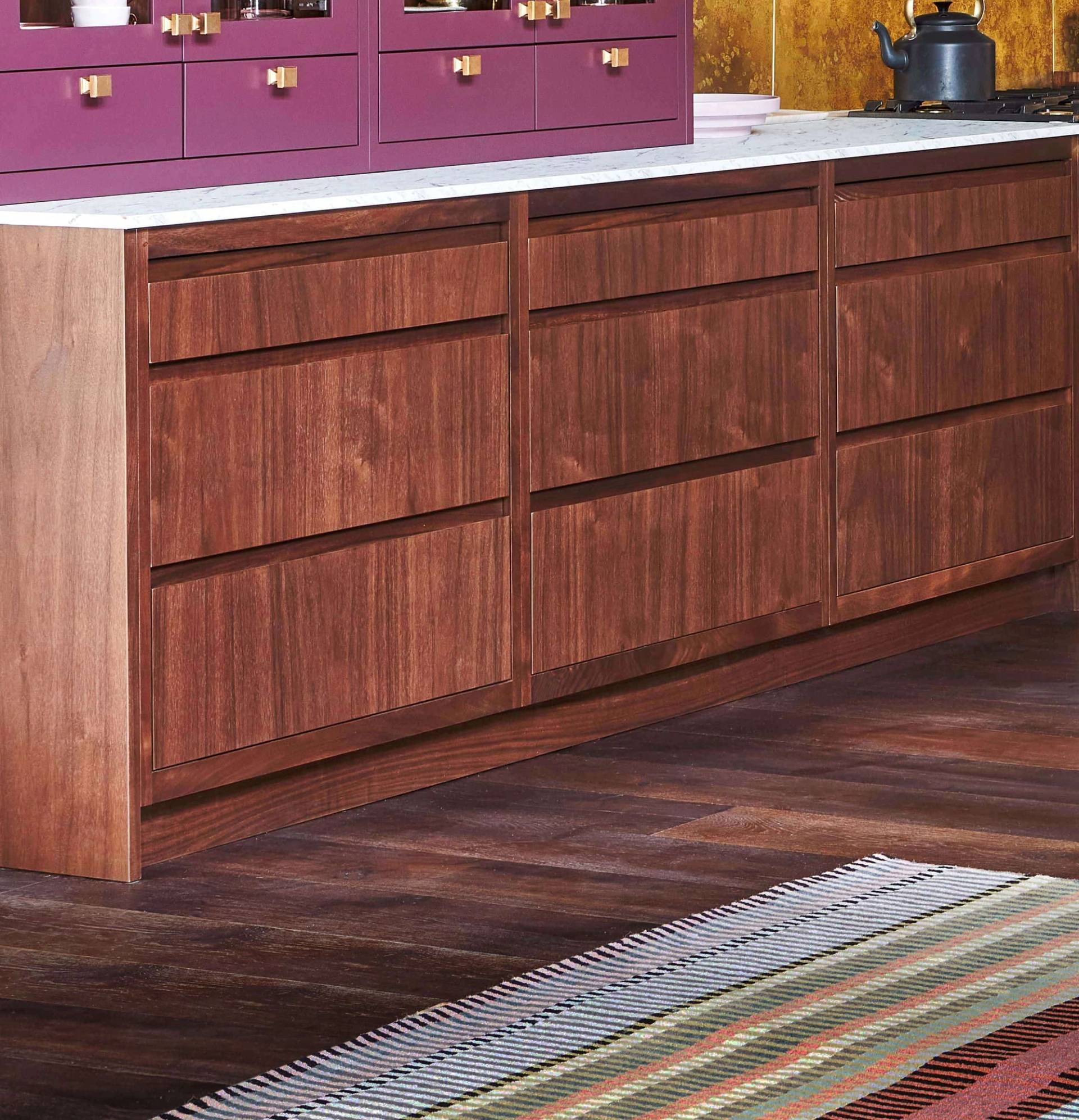 A close-up of a modern kitchen cabinet with three sets of wooden drawers, a white countertop, and a glimpse of a striped rug on a dark wood floor. Upper cabinets in purple with gold handles are partially visible.