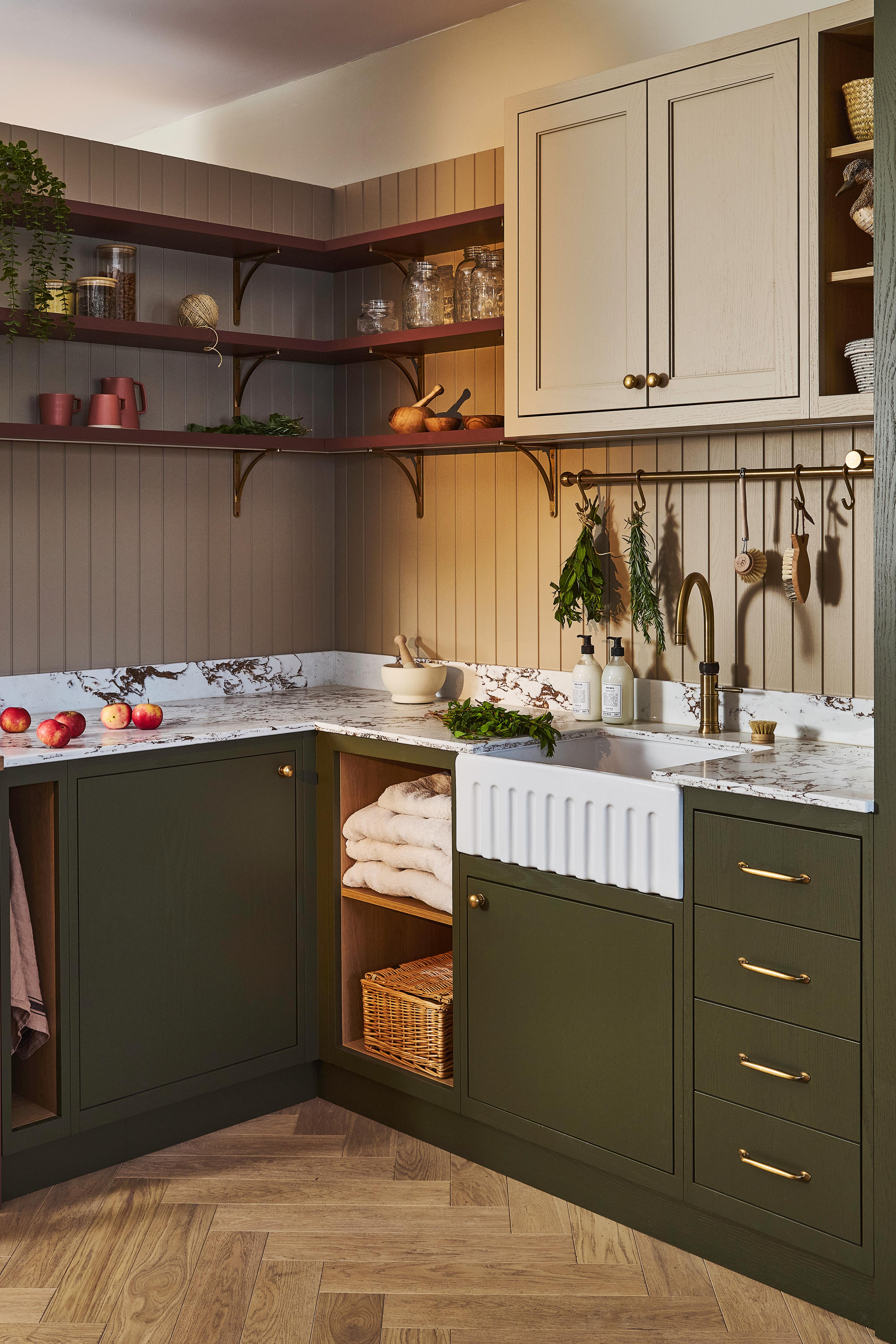 A stylish utility room with green cabinets, marble countertop, farmhouse sink, open shelves with dishes and plants, a washing machine behind a cabinet door, and neatly folded towels on a shelf.
