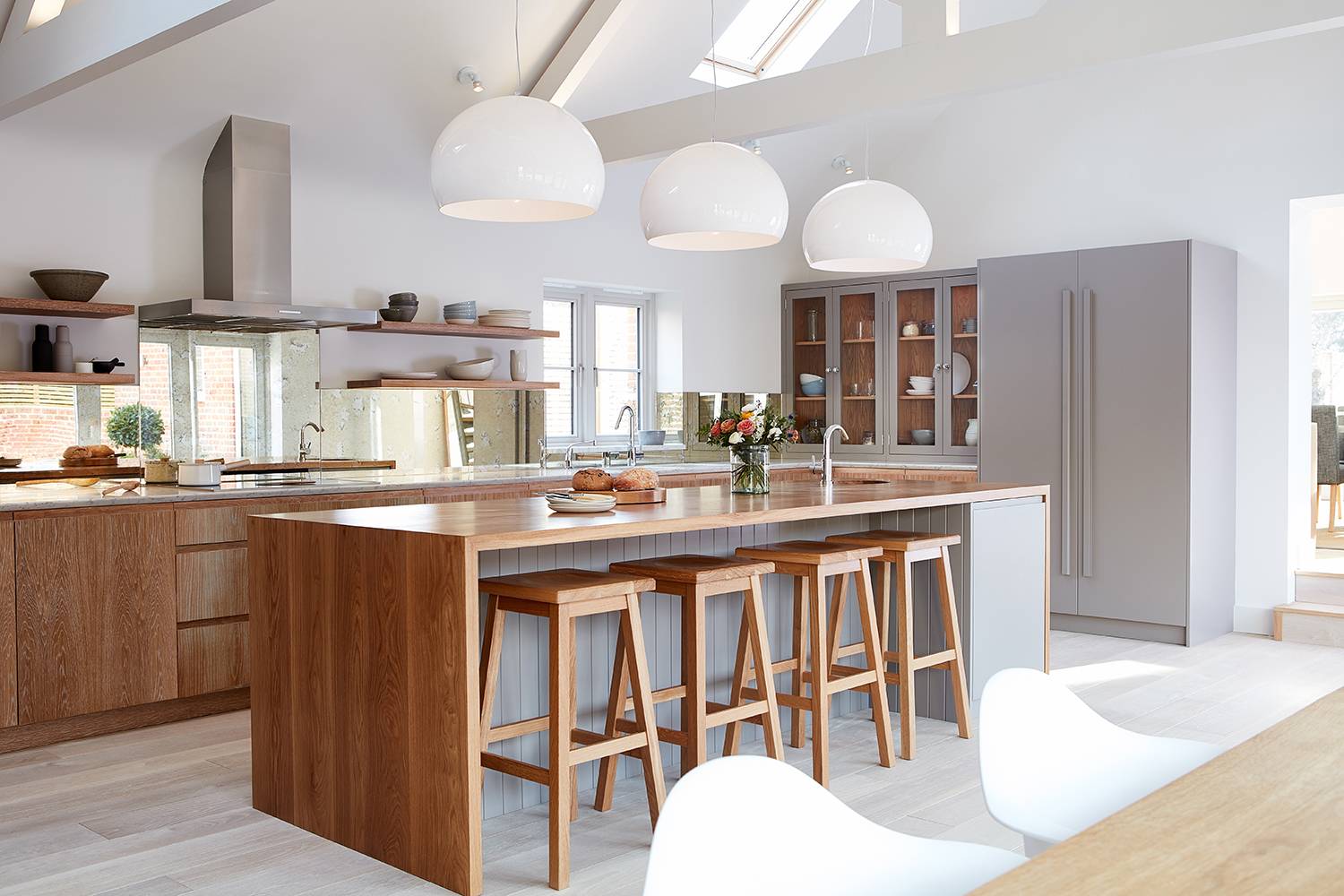 A modern kitchen with a large wooden island featuring four wooden stools. There are three large white pendant lights above. The kitchen has wooden cabinets, a grey refrigerator, and a built-in oven. Light floods in through skylights and windows.