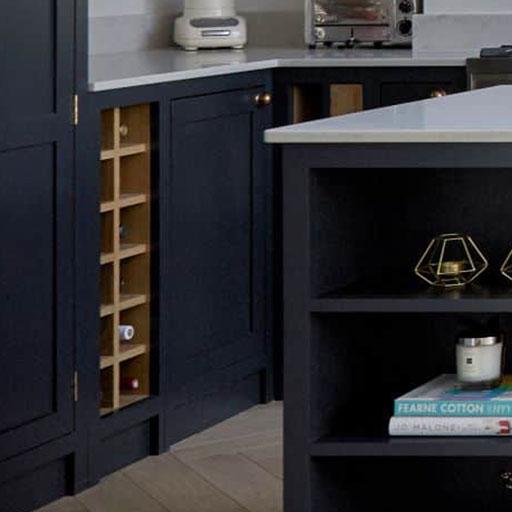 A modern kitchen corner with dark cabinets, a built-in wine rack, white countertop, and appliances. An island with shelves holds books, a candle, and geometric decor.