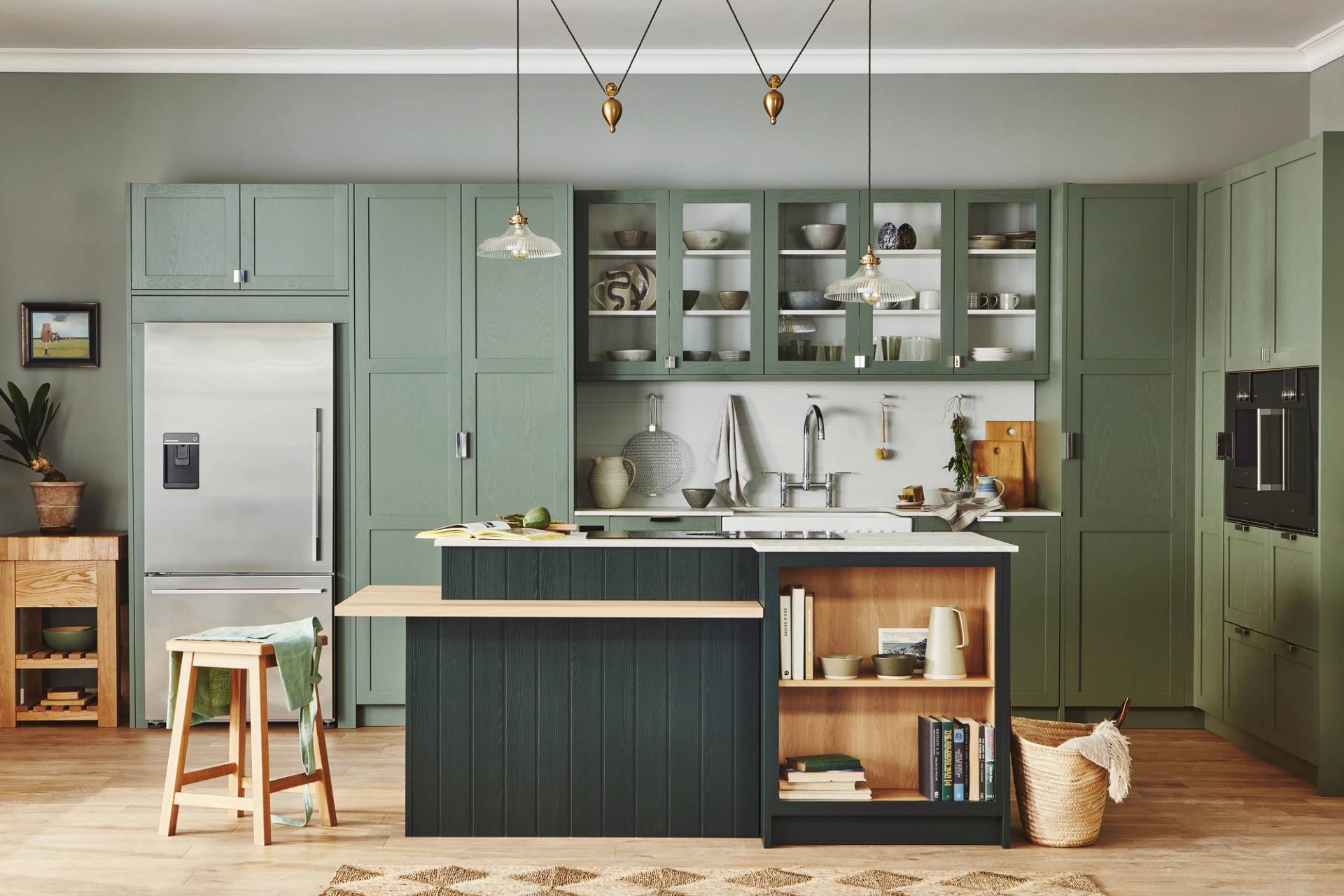 Modern kitchen with sage green cabinets, stainless steel appliances, open shelves displaying dishes, a center island with books and decor, a wooden stool, woven baskets, and pendant lights, all in a bright, tidy space.