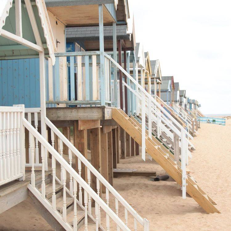 A row of colorful beach huts on a sandy beach, each with wooden stairs leading up to them. The image shows a clear sky and calm setting, with the huts slightly elevated above the sand.