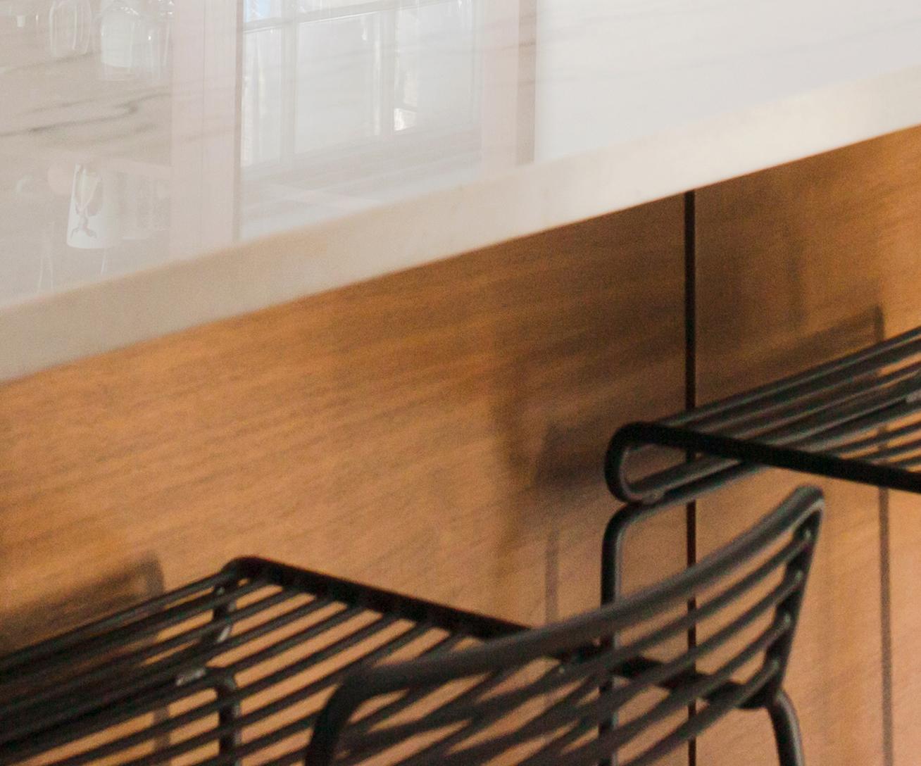 Close-up of a modern kitchen counter with a white marble surface and wooden front, featuring two black metal bar stools with slatted seats and backs.