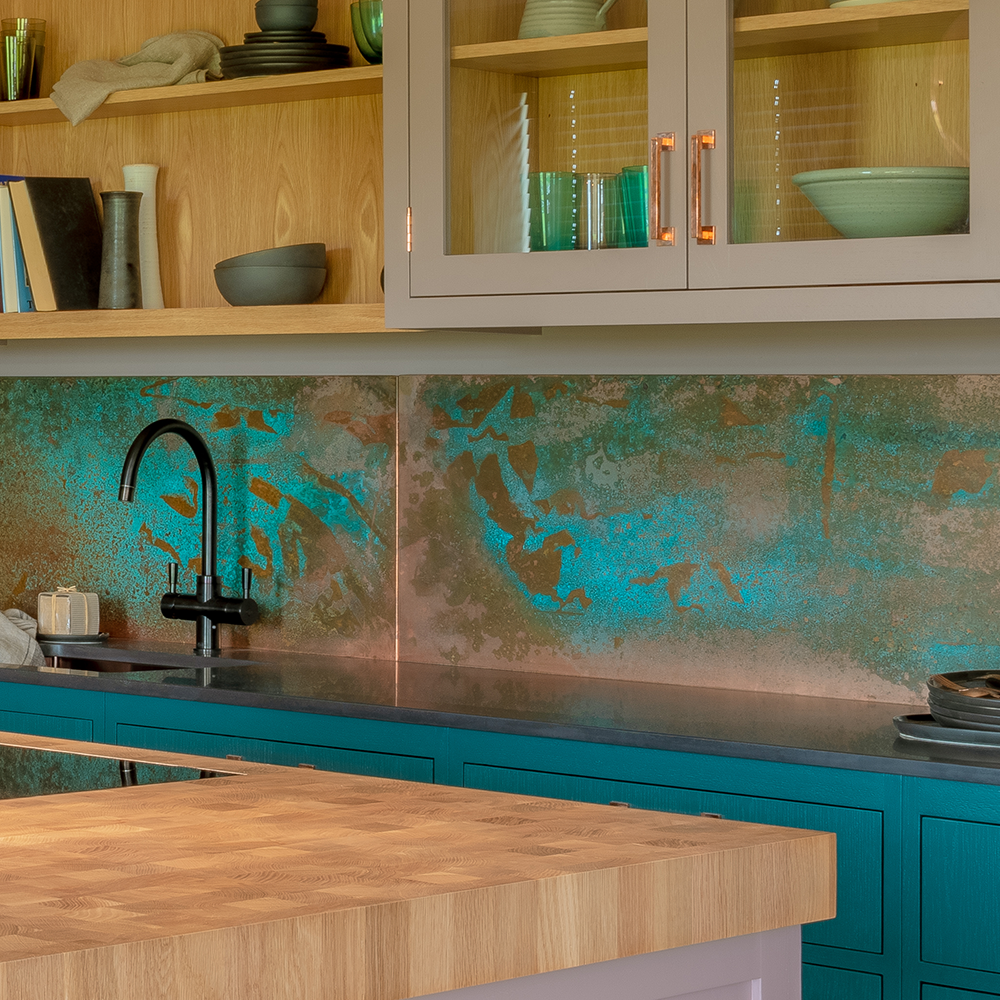 A modern kitchen with wooden countertops, teal cabinets, a black faucet, and a turquoise textured backsplash. Open shelves and glass-front cabinets display dishes and books.
