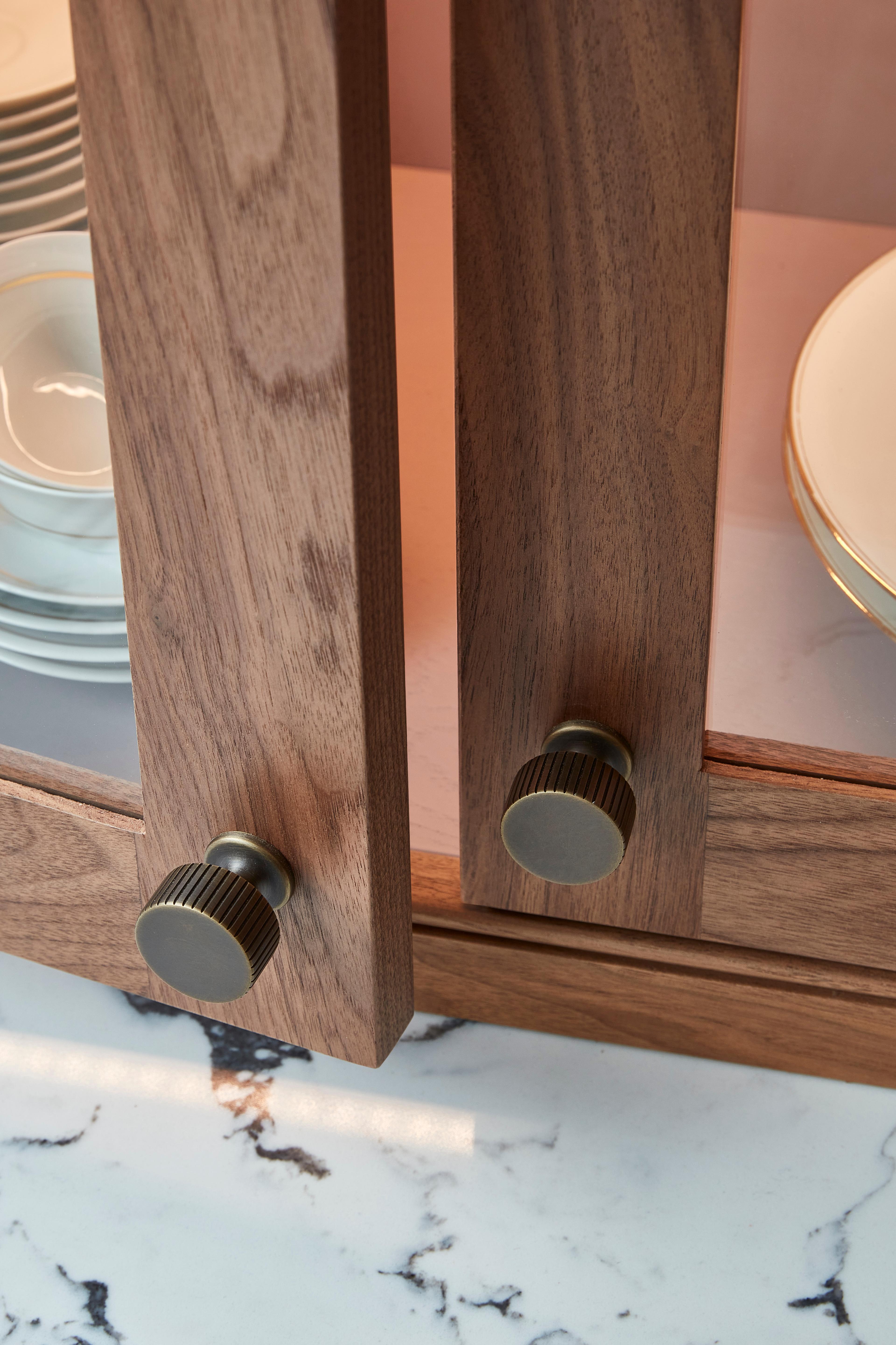 Close-up of a wooden cabinet with two open doors featuring round, textured metal knobs. Inside, there are neatly stacked plates with gold rims. The cabinet is set on a marbled surface