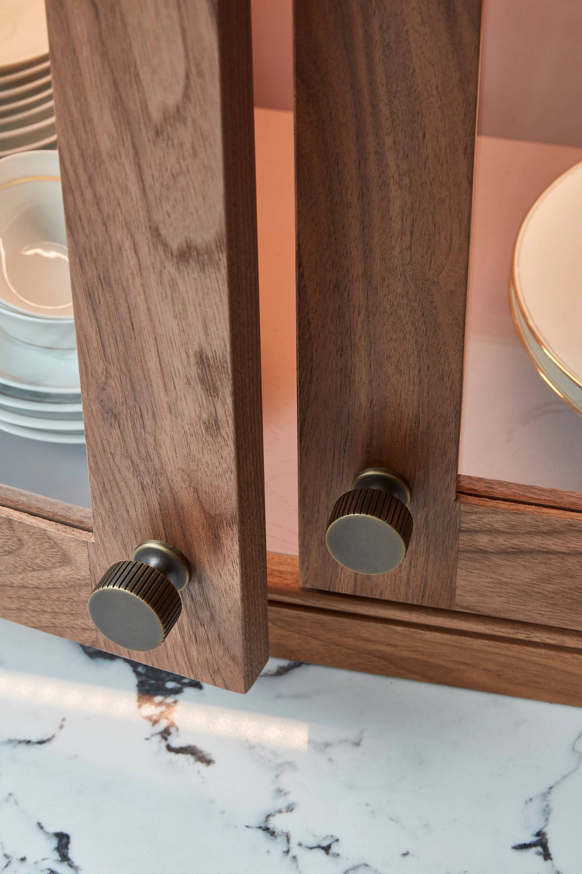 Close-up of a wooden cabinet with two open doors featuring round, textured metal knobs. Inside, there are neatly stacked plates with gold rims. The cabinet is set on a marbled surface