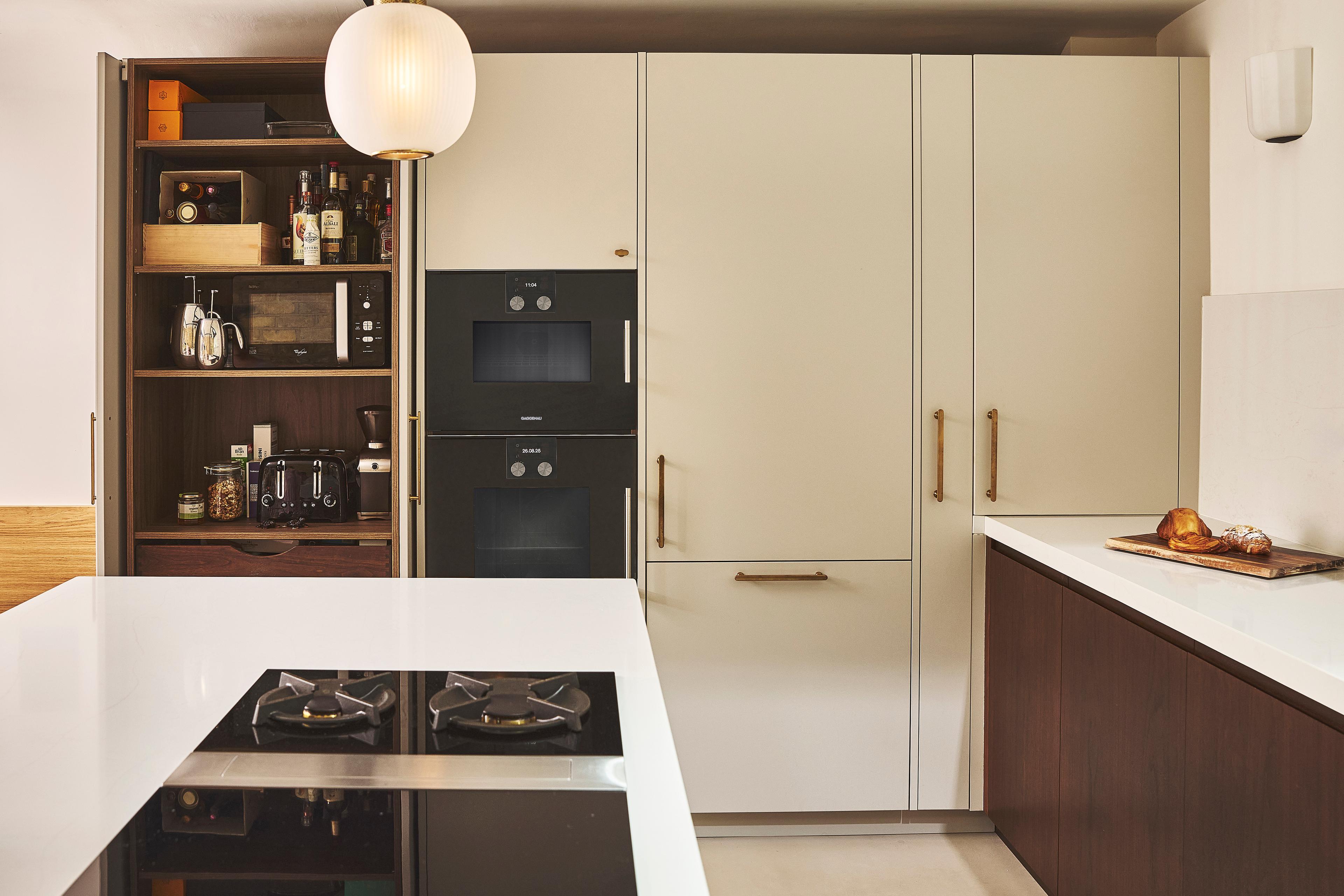 Modern kitchen with open white cabinets, built-in black oven, gold handles, a gas stove on a white island, and a countertop with a cutting board and bread. A round pendant light hangs from the ceiling