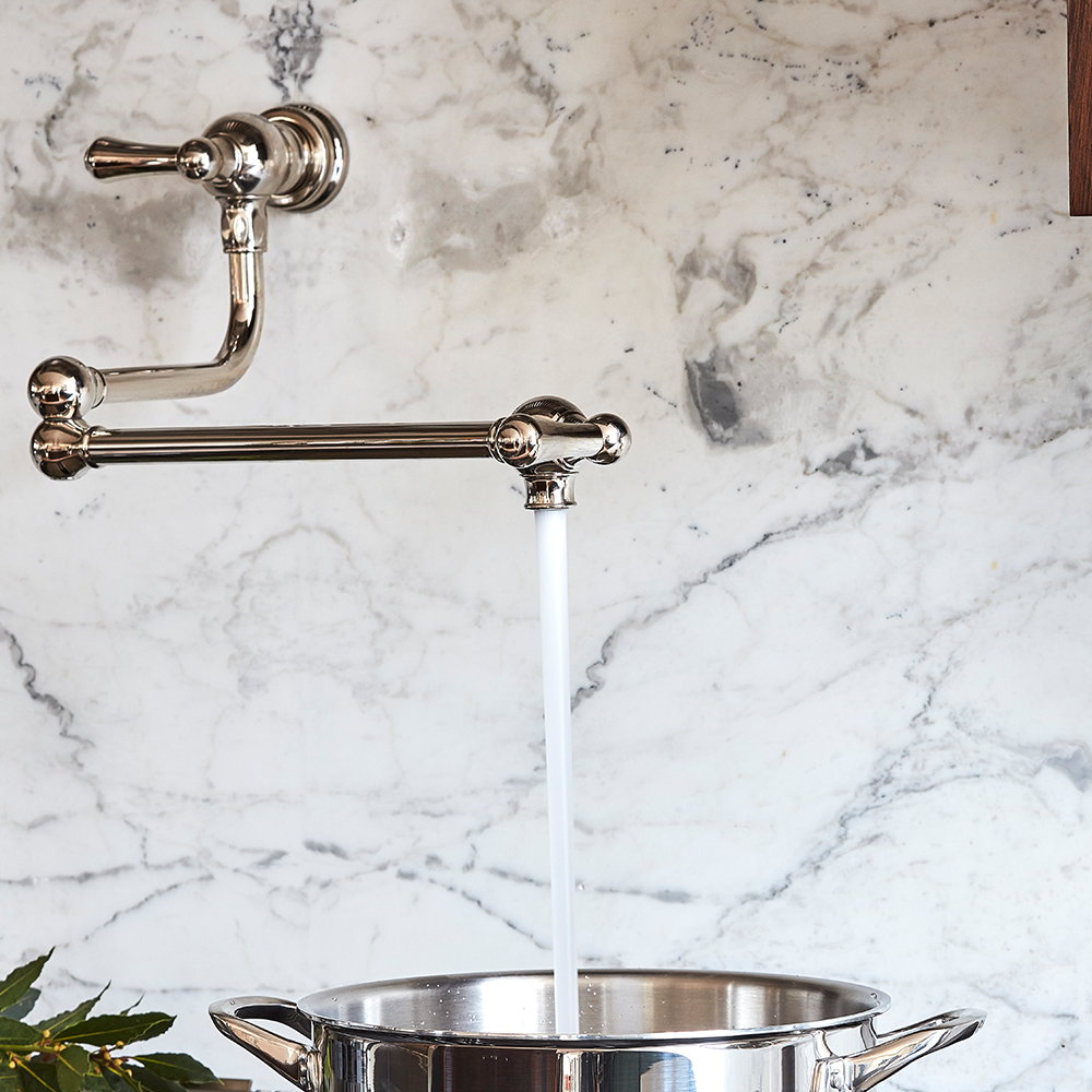A pot filler faucet attached to a marble backsplash releases a stream of water into a metal pot beneath it. Some green leaves are visible in the bottom left corner.