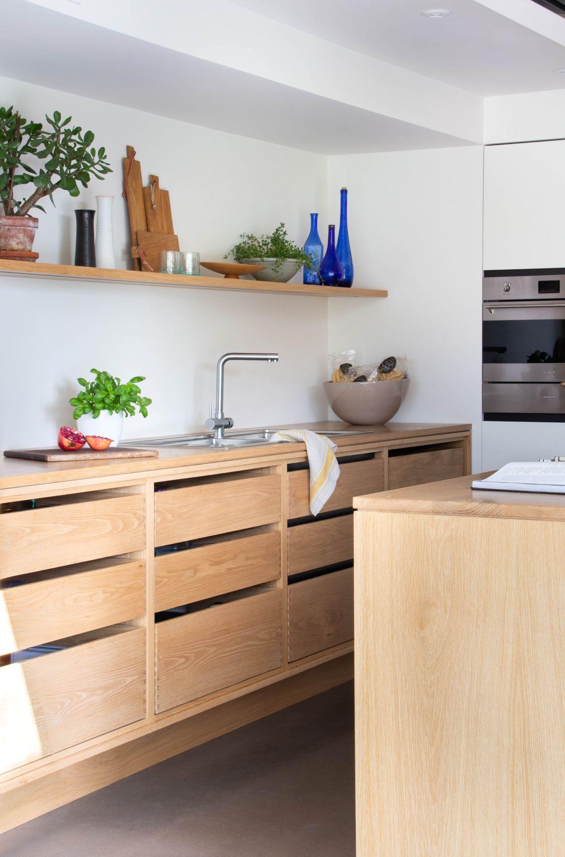A modern kitchen with wooden cabinets, a metal sink, and a countertop. A shelf above holds blue glass bottles, plants, and kitchen tools. A bowl of fruit is beside the sink. A stainless steel oven is built into the wall.