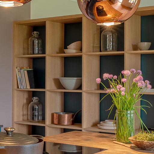 Open wooden shelving unit with dishes, books, and glass jars against a dark backing. A glass vase with purple flowers sits on a wooden kitchen counter in the foreground, with copper pendant lights hanging above.