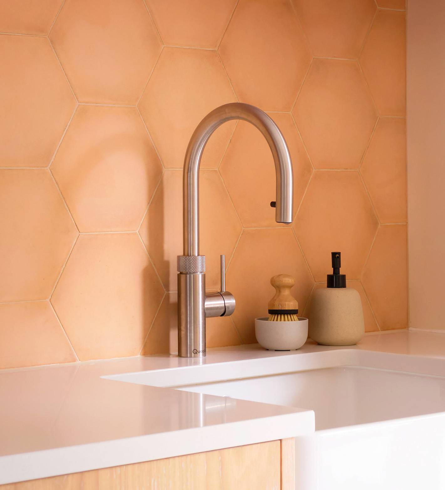 A modern kitchen sink with a sleek stainless steel faucet, set against a peach hexagon tile backsplash. On the counter are a small dish brush and a soap dispenser