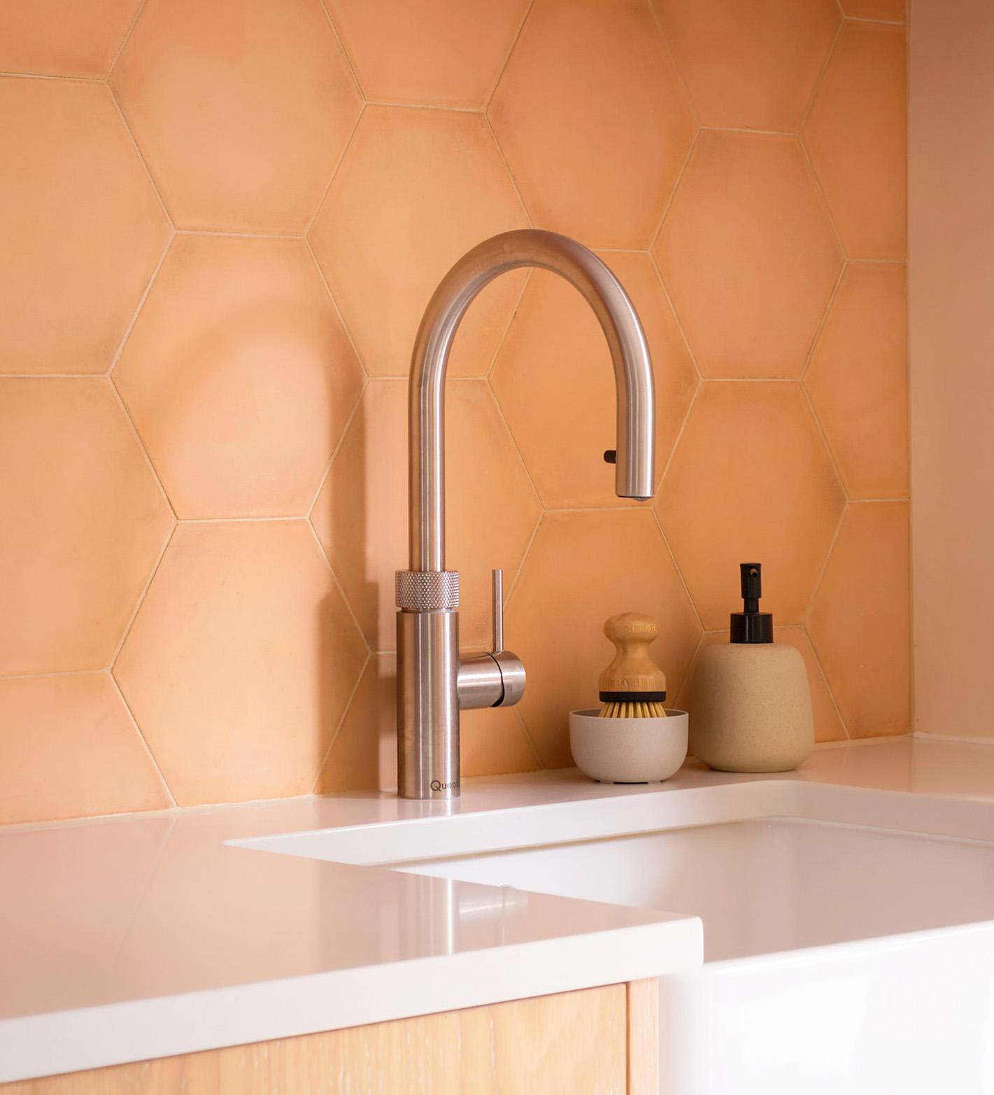 A modern kitchen sink with a sleek stainless steel faucet, set against a peach hexagon tile backsplash. On the counter are a small dish brush and a soap dispenser
