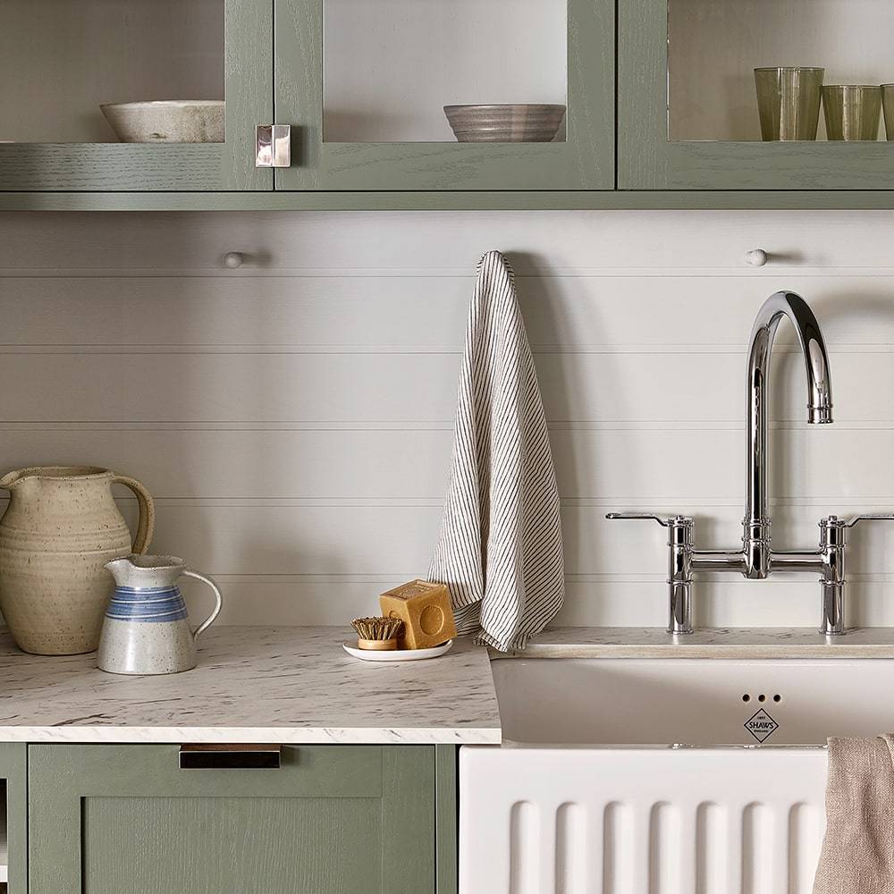 A kitchen with green cabinets and a white countertop. A striped towel hangs near a silver faucet over a farmhouse sink. Ceramic jugs, stacked bowls, and glasses are placed on the counter and shelves.