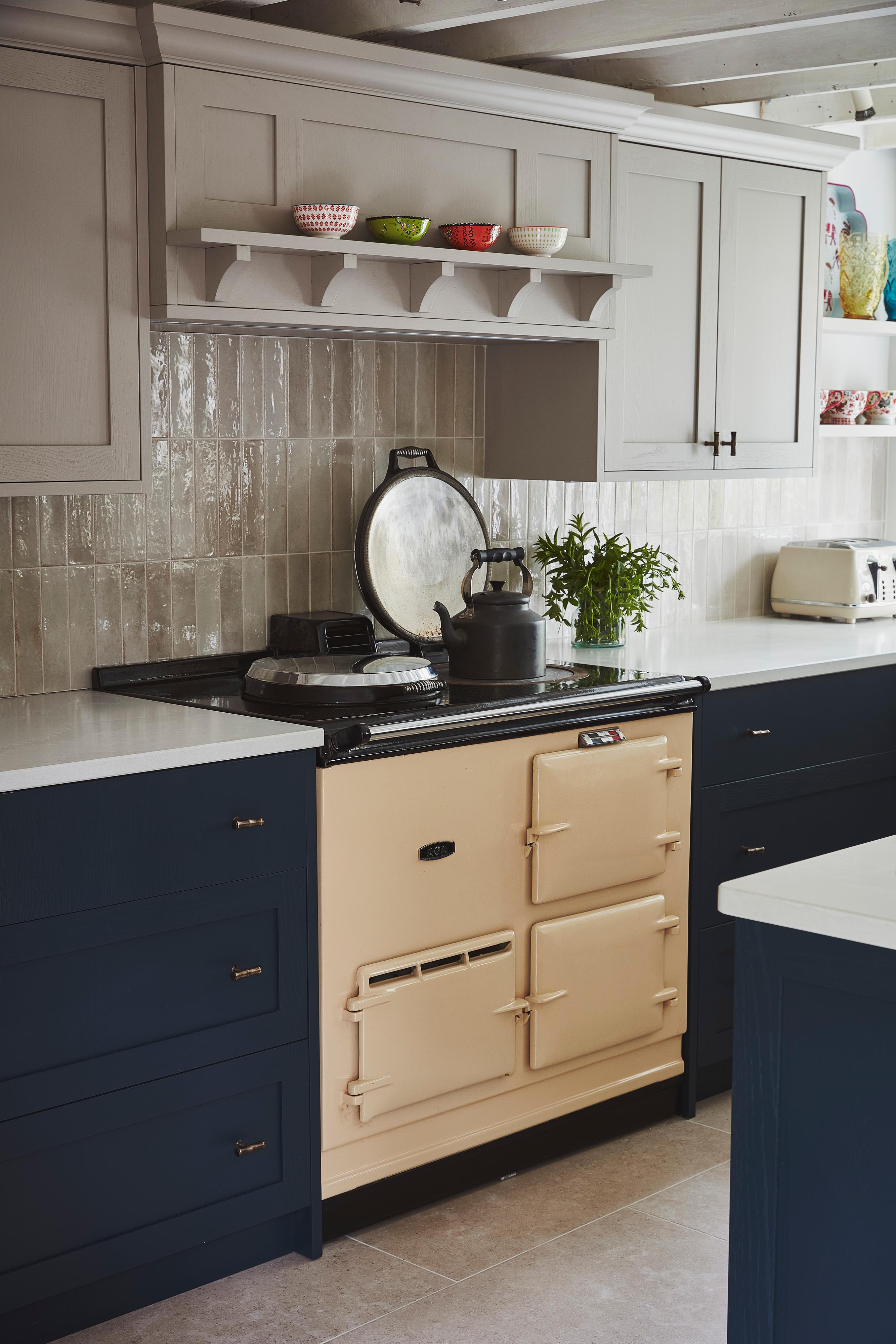 A cozy kitchen with navy blue and light gray cabinets, cream-colored vintage stove, white countertops, tiled backsplash, and shelves with colorful bowls. A kettle and a pot are on the stove with herbs nearby.