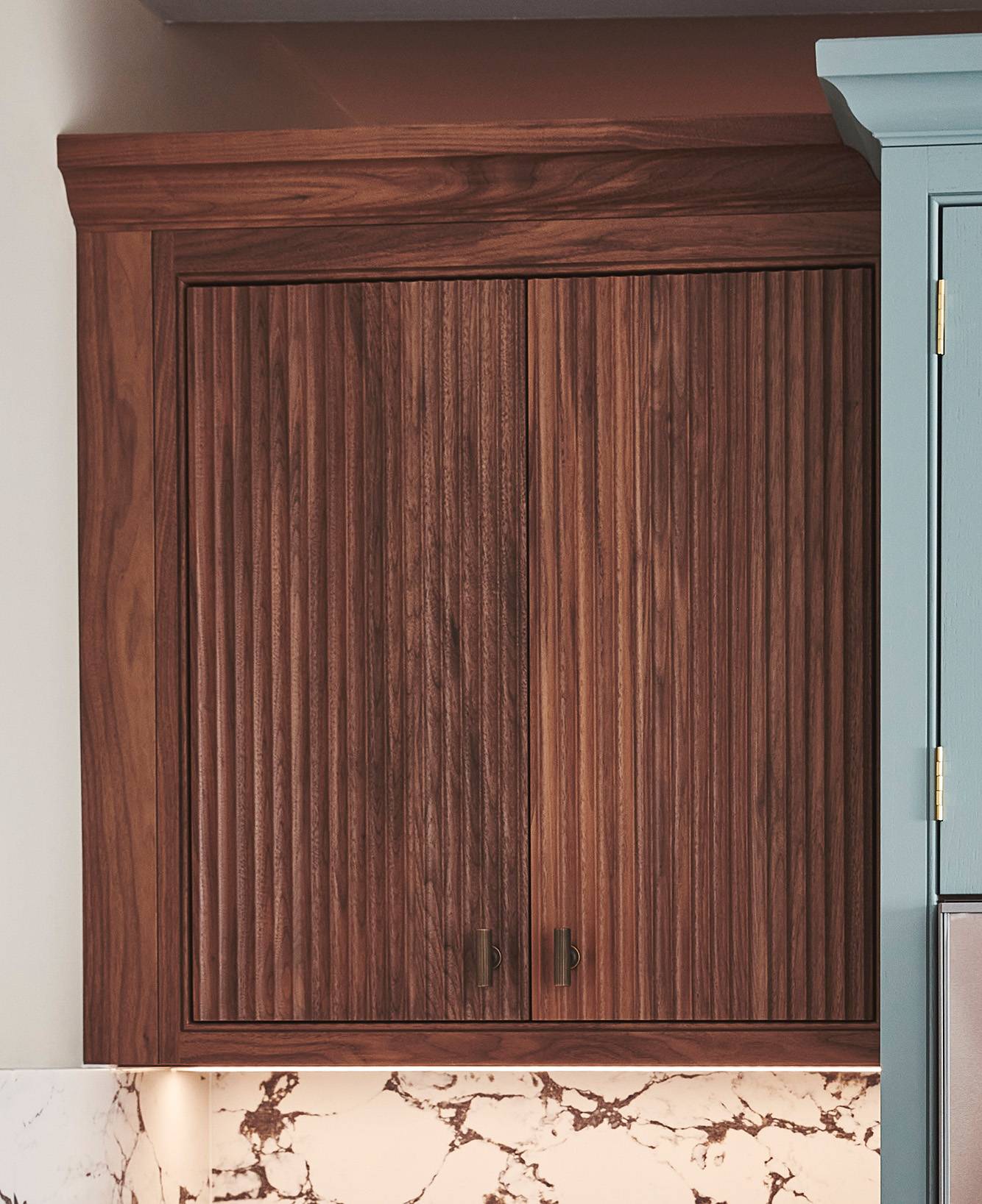 Wooden kitchen cabinet with vertical grooves and two metal handles, set against a wall. The bottom of the image shows part of a white and brown marble countertop.