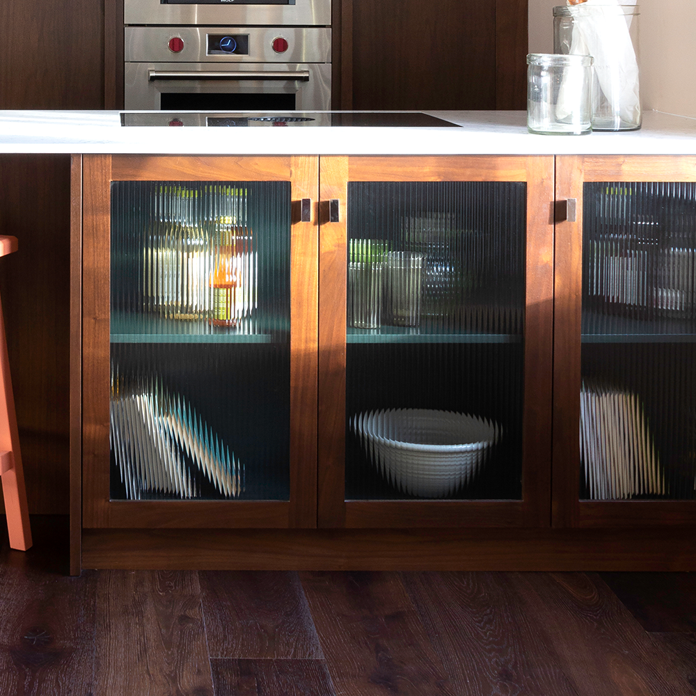 A kitchen with a dark wood cabinet featuring glass doors. Inside are jars, bottles, and a white bowl. The countertop above is white, and kitchen appliances are visible in the background.