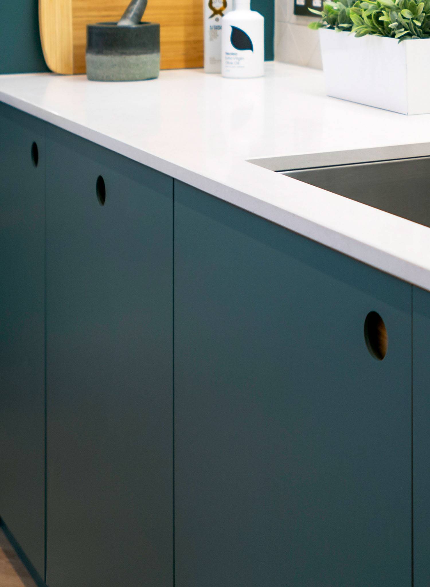 A modern kitchen counter with matte dark teal cabinets, a white countertop, a built-in sink, a white planter with green leaves, a cutting board, and a jar.