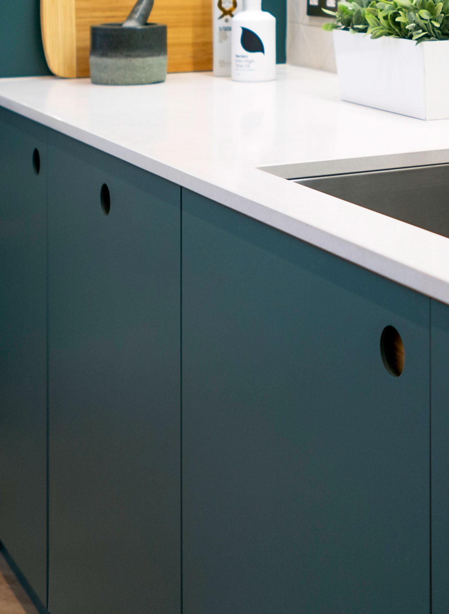 A modern kitchen counter with matte dark teal cabinets, a white countertop, a built-in sink, a white planter with green leaves, a cutting board, and a jar.