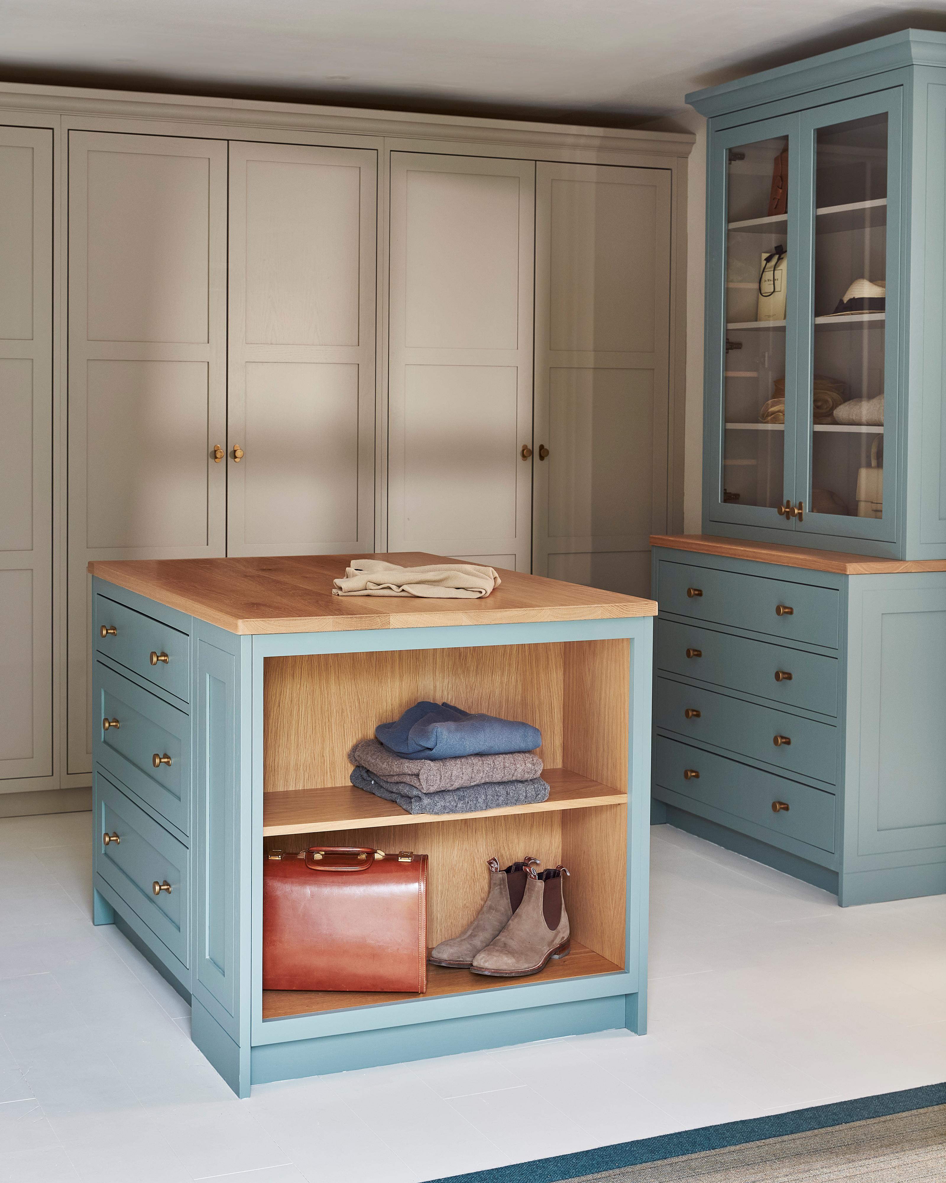 A spacious, organized walk-in closet with light blue built-in cabinets, a wood-topped island with open shelves holding folded clothes, boots, and a leather bag, and floor-to-ceiling gray wardrobes in the background.