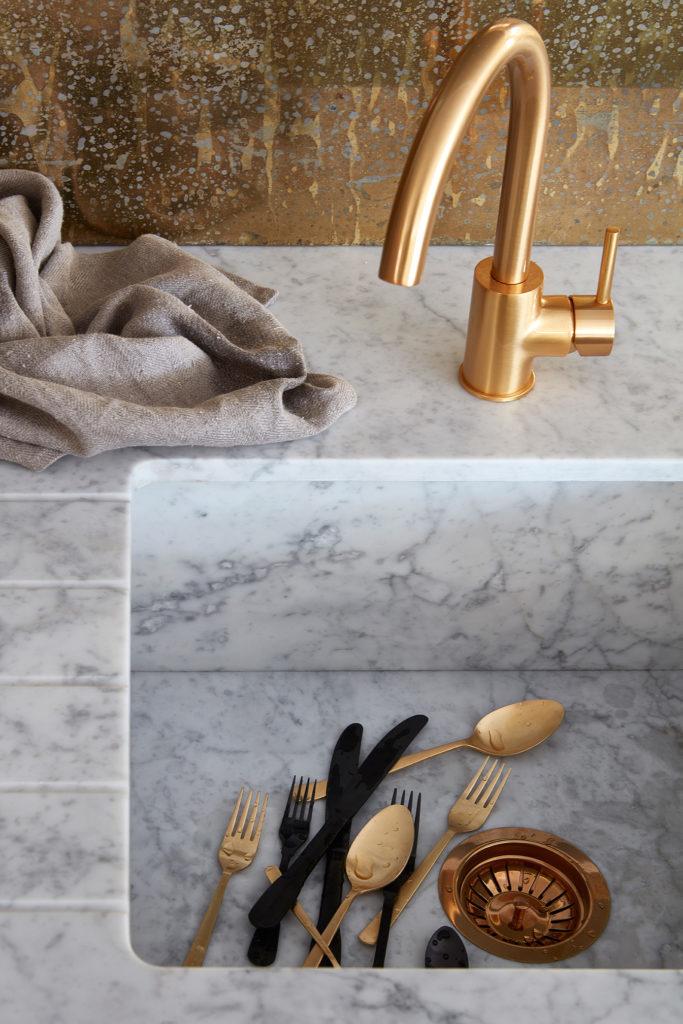 Close-up of an integrated white marble sink with grooved drainboard, featuring a brushed brass swan-neck tap and aged brass backsplash wall.