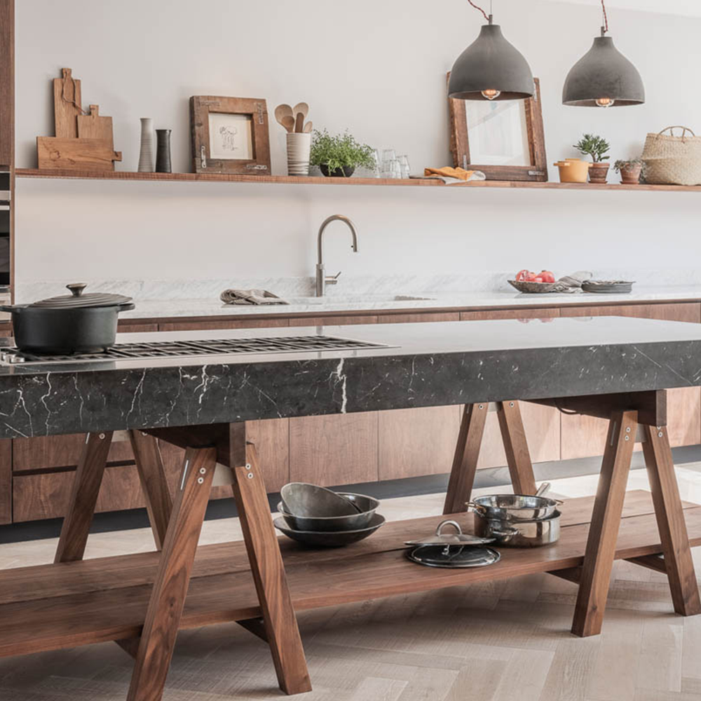 A modern kitchen featuring a dark marble-topped island with wooden legs. Pots and pans are stored underneath. Brown cabinets line the back wall, with a sink and various decor items. Two black pendant lights hang overhead.