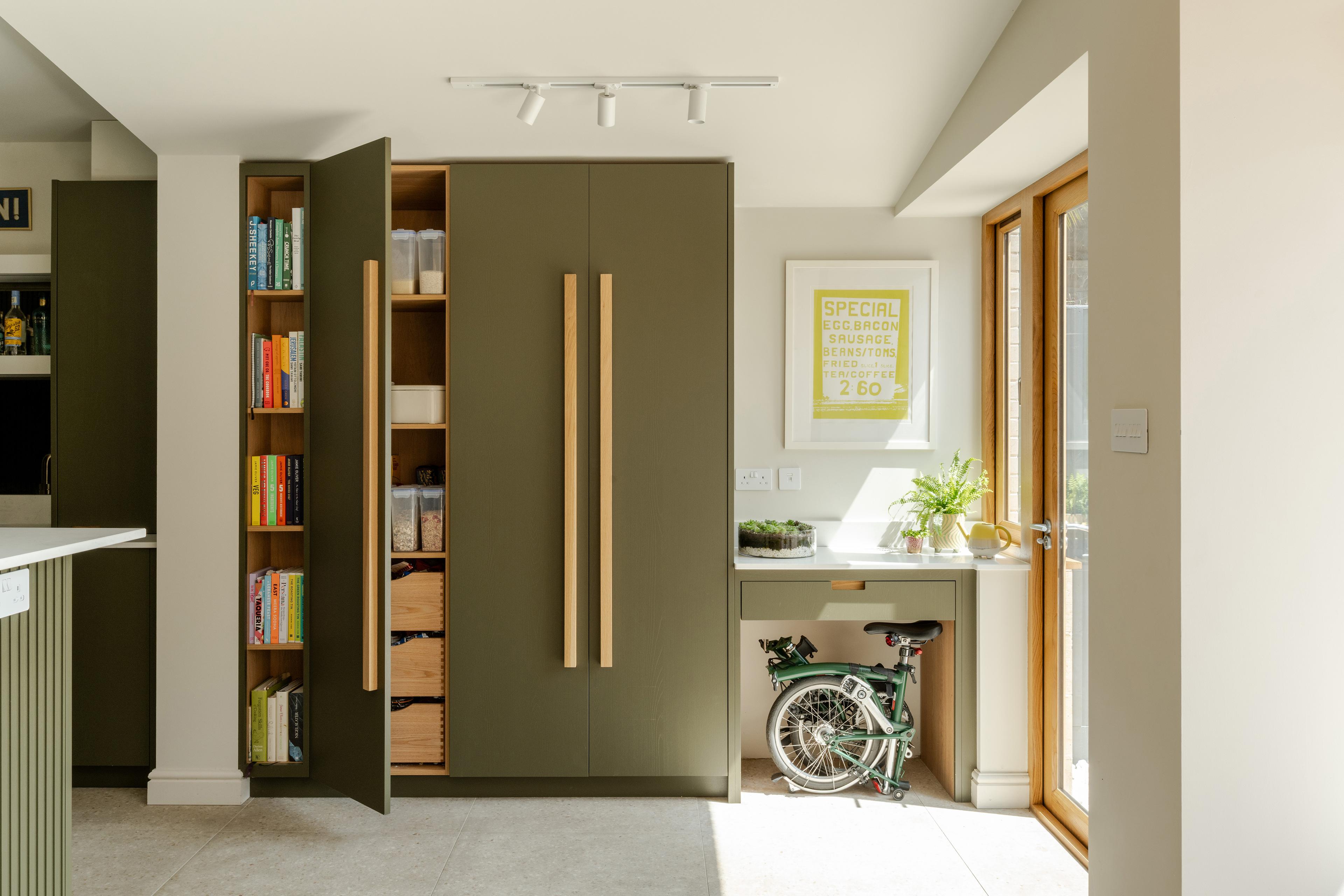 A modern kitchen with olive-green cabinets, open shelving with books and baskets, a small folding bicycle stored under a desk, and sunlight streaming through a glass door. A framed poster and potted plant are on the desk.