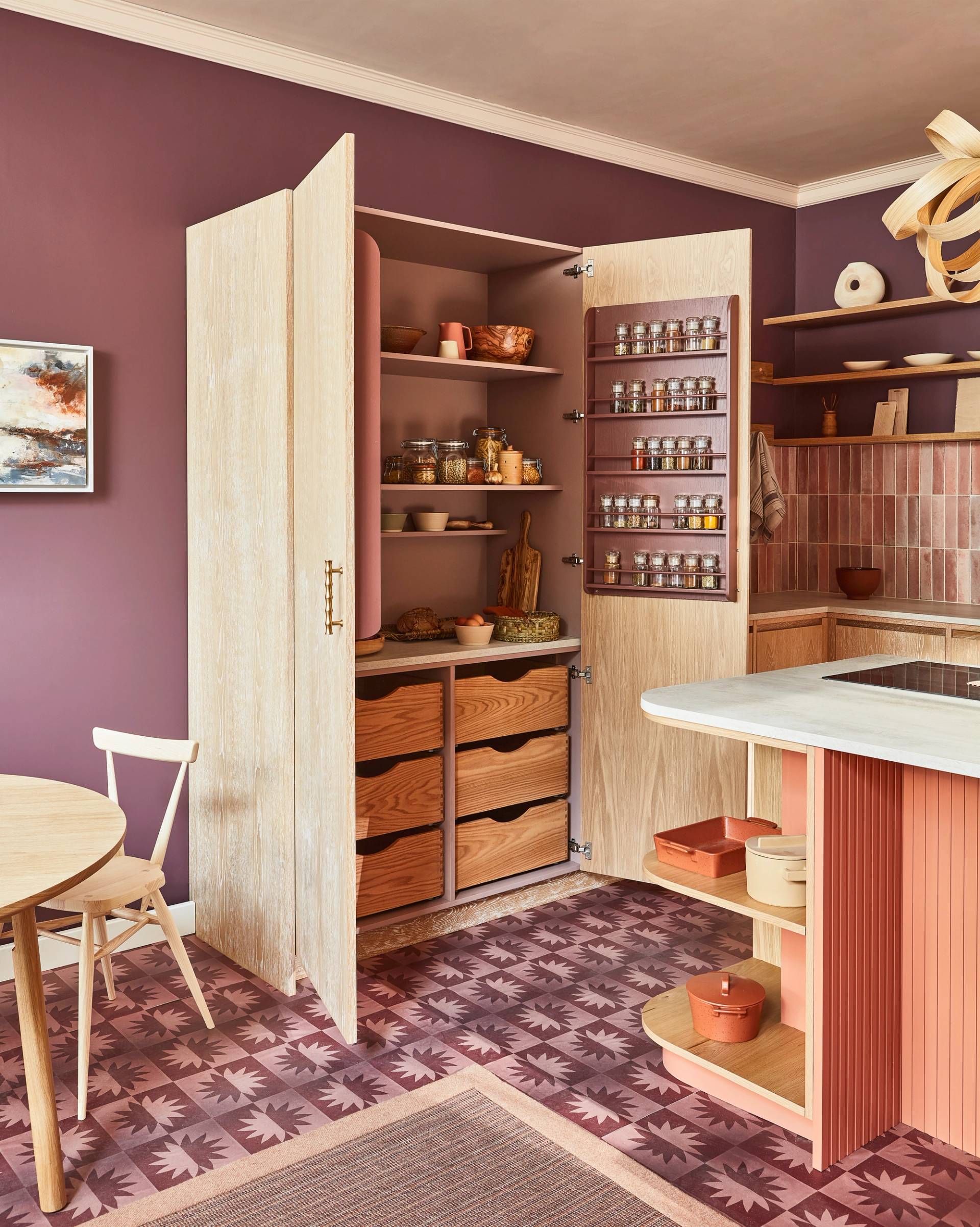 A stylish kitchen with purple walls and a patterned floor. A wooden cabinet is open, revealing shelves stocked with jars and kitchenware. A round table with a chair is nearby, creating a cozy and organized culinary space.