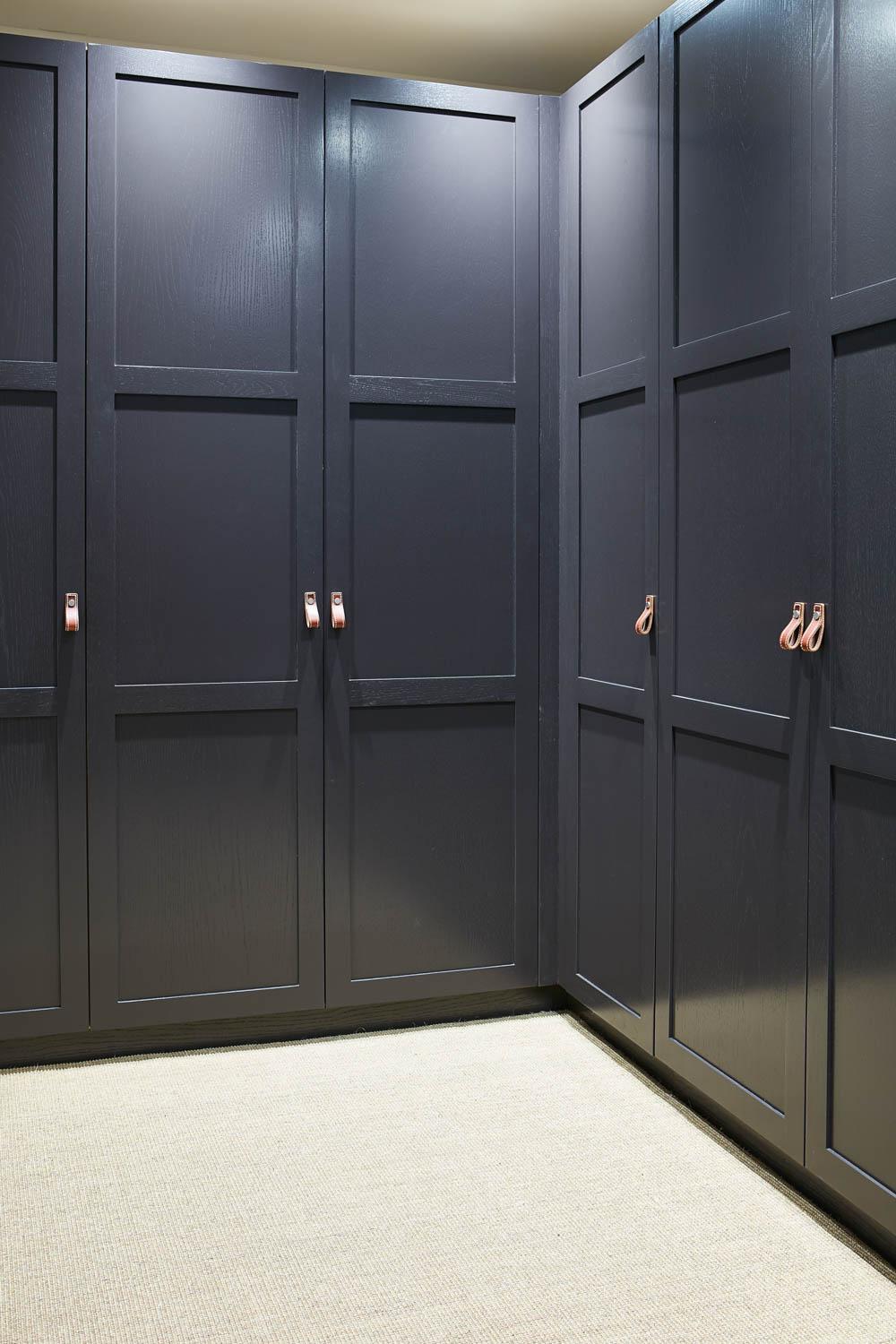 A modern dark wood wardrobe with one door open, revealing wooden drawers, neatly folded clothes, and a hanging jacket inside. The wardrobe stands on a light-colored carpeted floor