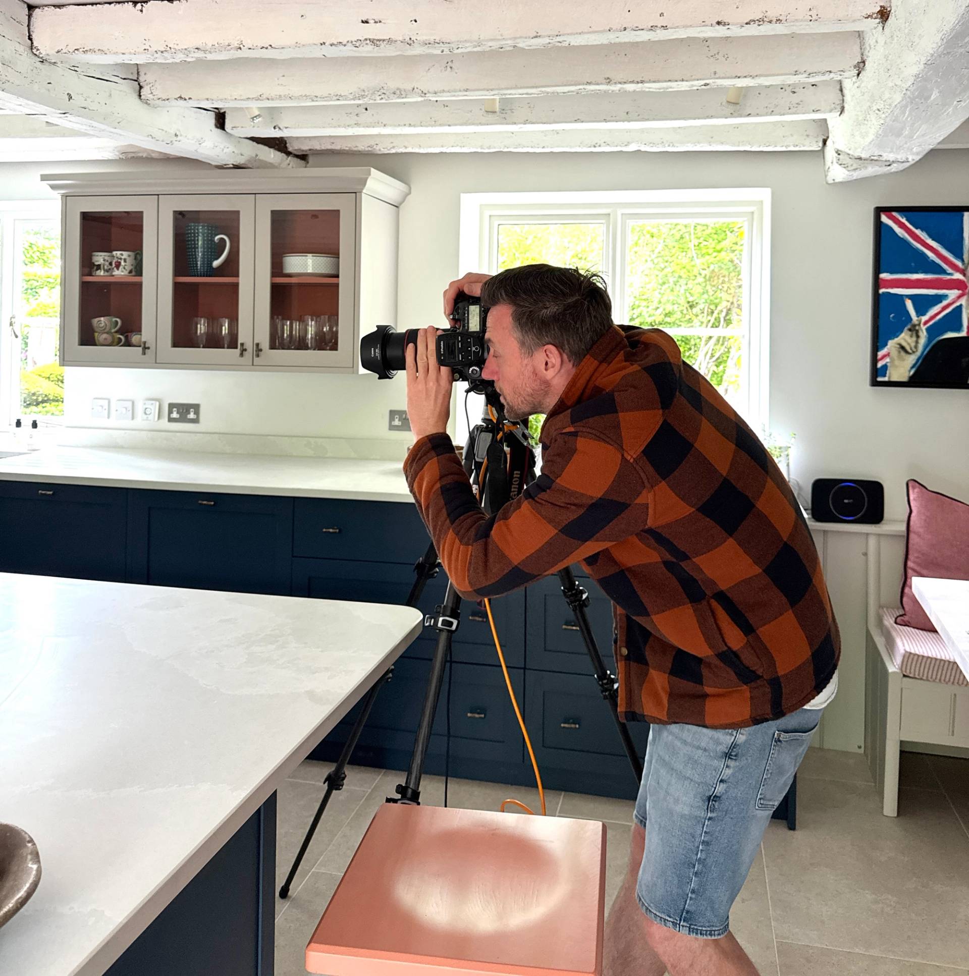 A man in a checkered shirt uses a professional camera on a tripod to photograph a modern kitchen with blue cabinets.