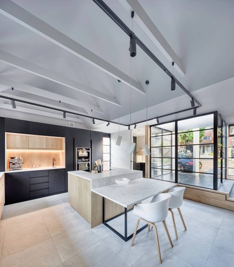 Modern kitchen with high ceilings and exposed beams. Features a central island with white chairs, marble countertops, sleek black and wood cabinetry, and large windows allowing natural light. Minimalistic design with pendant lighting.