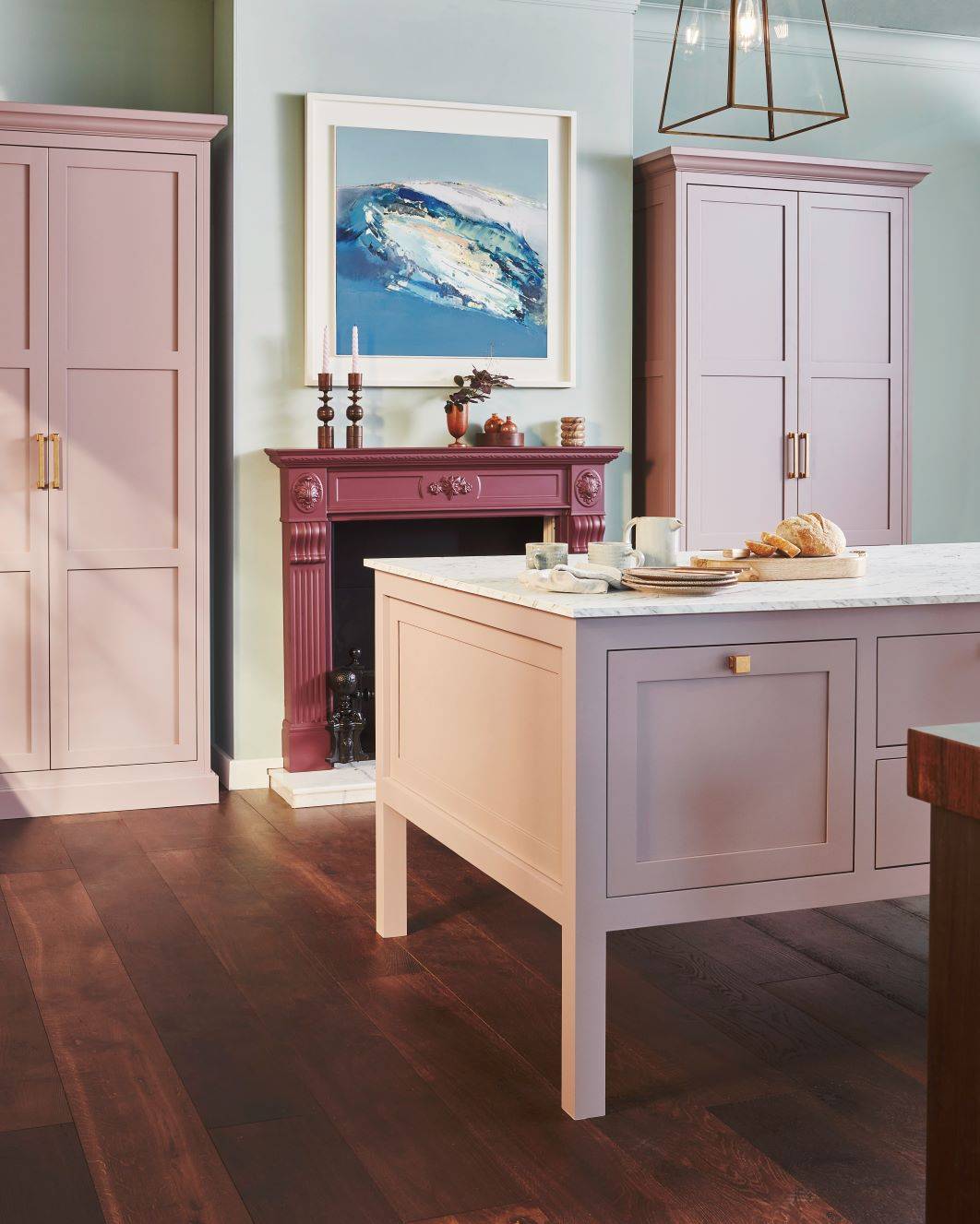 A stylish kitchen featuring pastel pink cabinets, a dark wood floor, and a countertop with bread and mugs. A framed abstract painting hangs above a decorative burgundy fireplace with candles and decor items on the mantel.