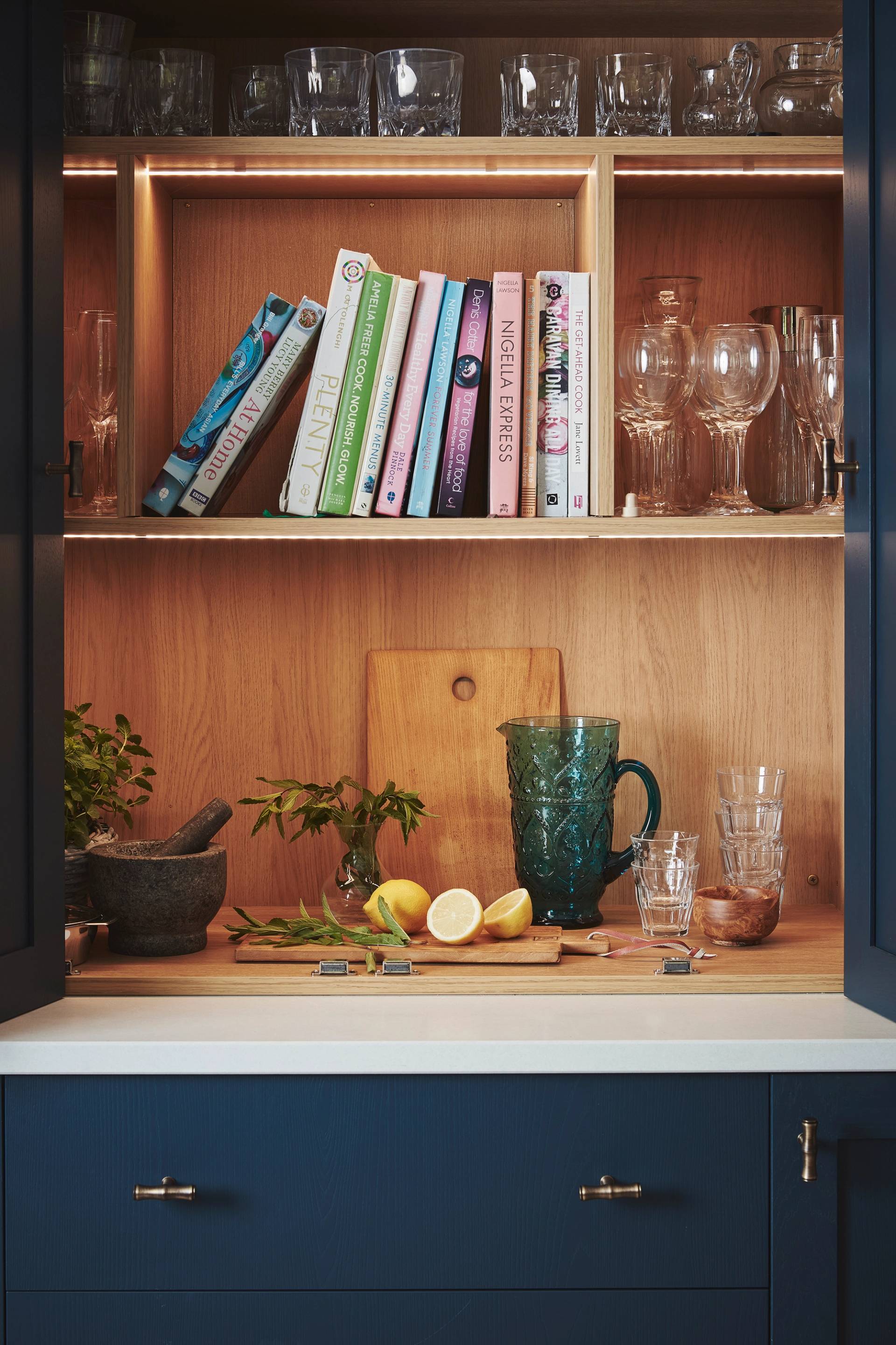 A kitchen cabinet with glassware, cookbooks, and a wooden cutting board. On the counter are a green glass pitcher, stacked glasses, a mortar and pestle, herbs, and a halved lemon with a knife.