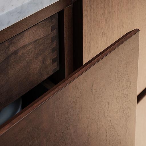 Close-up of a wooden cabinet with an open drawer, revealing dovetail joinery. The drawer has a smooth wood finish, and the countertop above appears to be light-colored, possibly marble or granite.