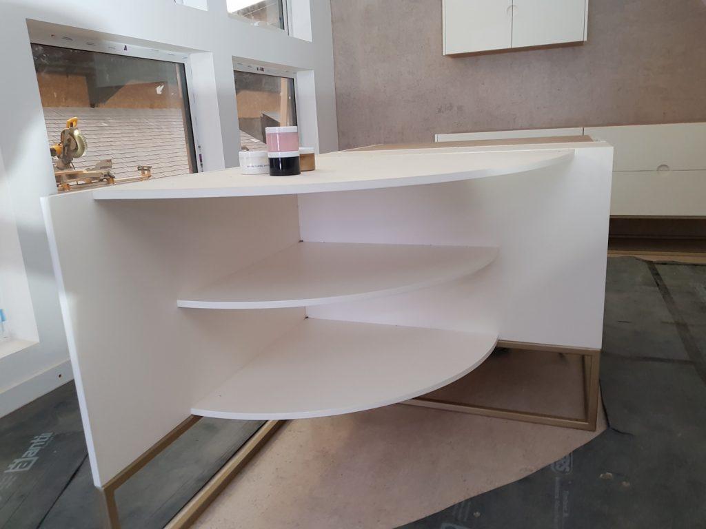 A modern white kitchen island unit under construction, featuring an overhanging curved countertop and matching open curved shelves supported by a brass-finished frame.