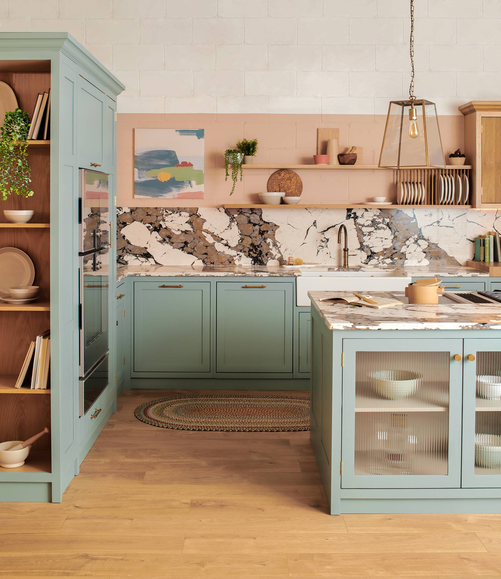 A modern kitchen and dining area with a marble island, wooden cabinets, and a built-in stovetop. The open layout features large windows, a green accent wall, and minimalist pendant lights over a dining table with white chairs