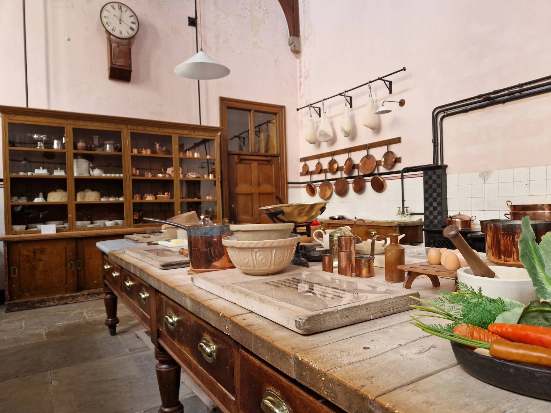 An eye-level shot of a historic Victorian kitchen prep table laden with antique ceramic bowls, brass measuring cups, and fresh vegetables, set against a background of large wooden display cabinets and copper pans hanging on pale pink walls.