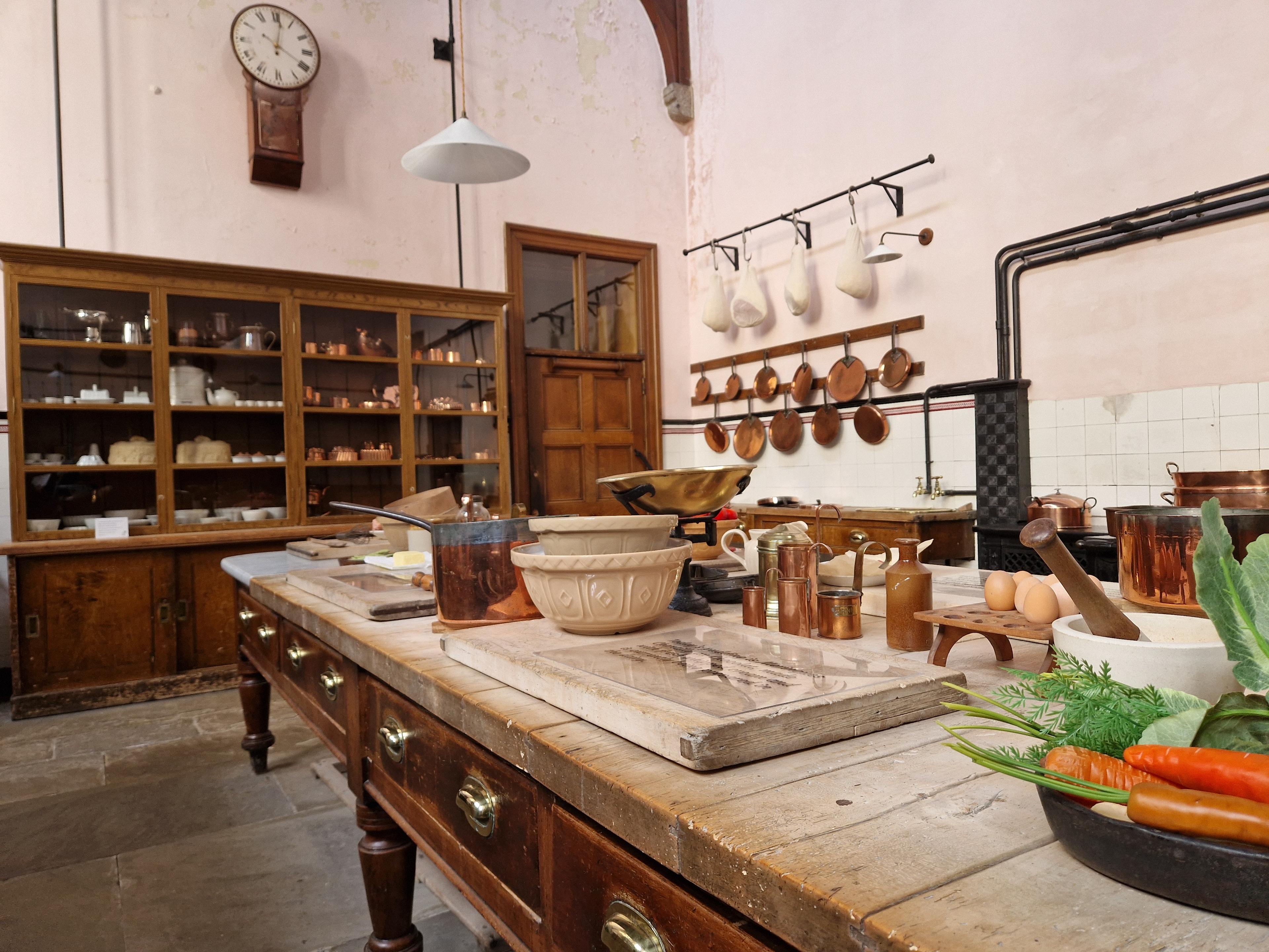 An eye-level shot of a historic Victorian kitchen prep table laden with antique ceramic bowls, brass measuring cups, and fresh vegetables, set against a background of large wooden display cabinets and copper pans hanging on pale pink walls.