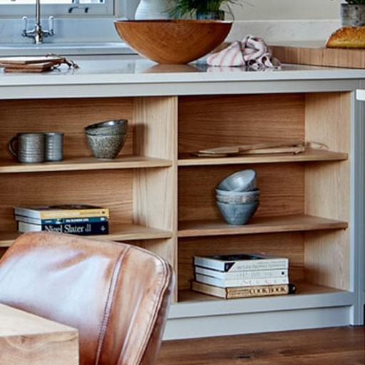 A kitchen island with open wooden shelves holding ceramic bowls, cups, a wooden tray, and stacked books. A countertop above displays a large wooden bowl, a loaf of bread, and a folded cloth near a window.