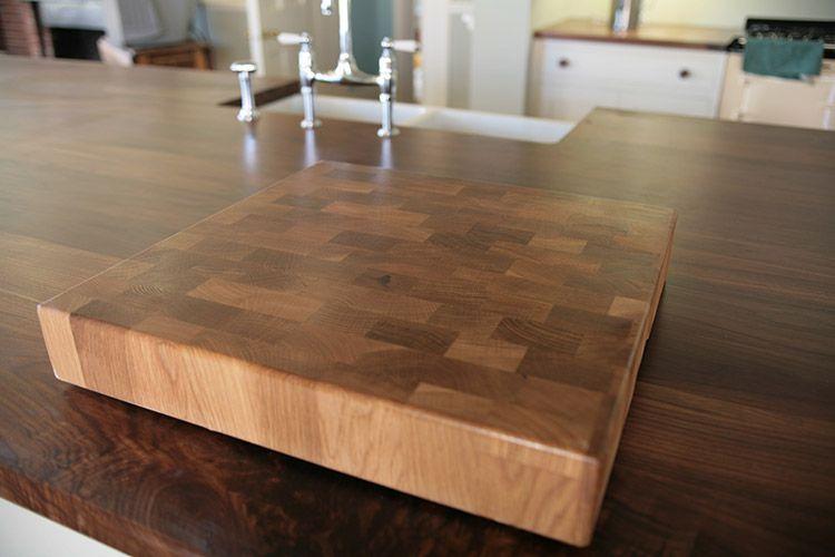 A large wooden cutting board with a checkerboard pattern sits on a smooth, dark wood kitchen countertop. A kitchen sink with a faucet is visible in the background, surrounded by light-colored cabinets.