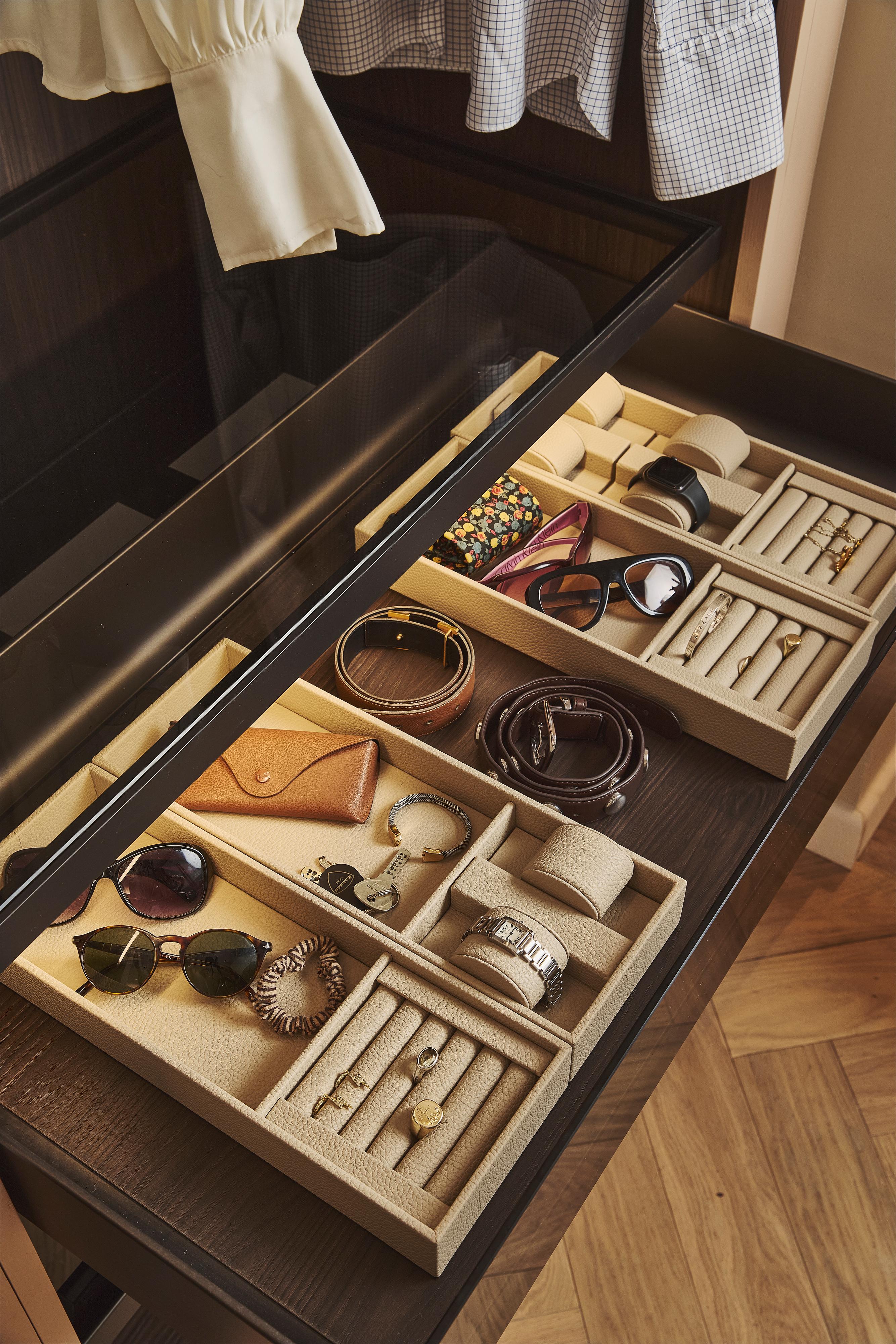 Open drawer with organized compartments holding sunglasses, watches, bracelets, rings, a hair tie, a jewelry pouch, and other small accessories. Clothes are hanging above the drawer. The drawer interior is wooden.