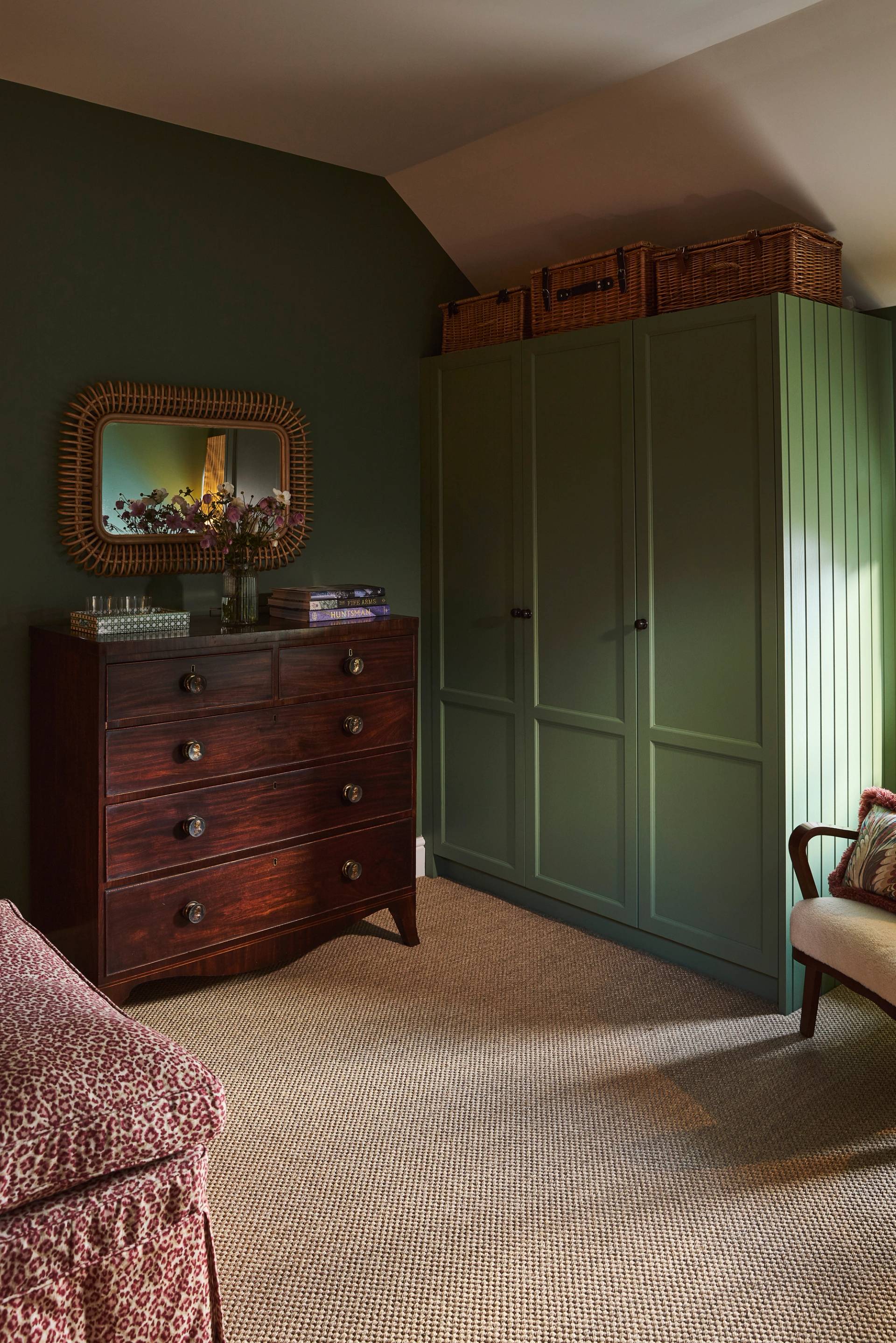 A cozy bedroom corner with olive green walls, a matching green wardrobe with baskets on top, a dark wood chest of drawers with a decorative mirror above, and a patterned armchair and carpet.
