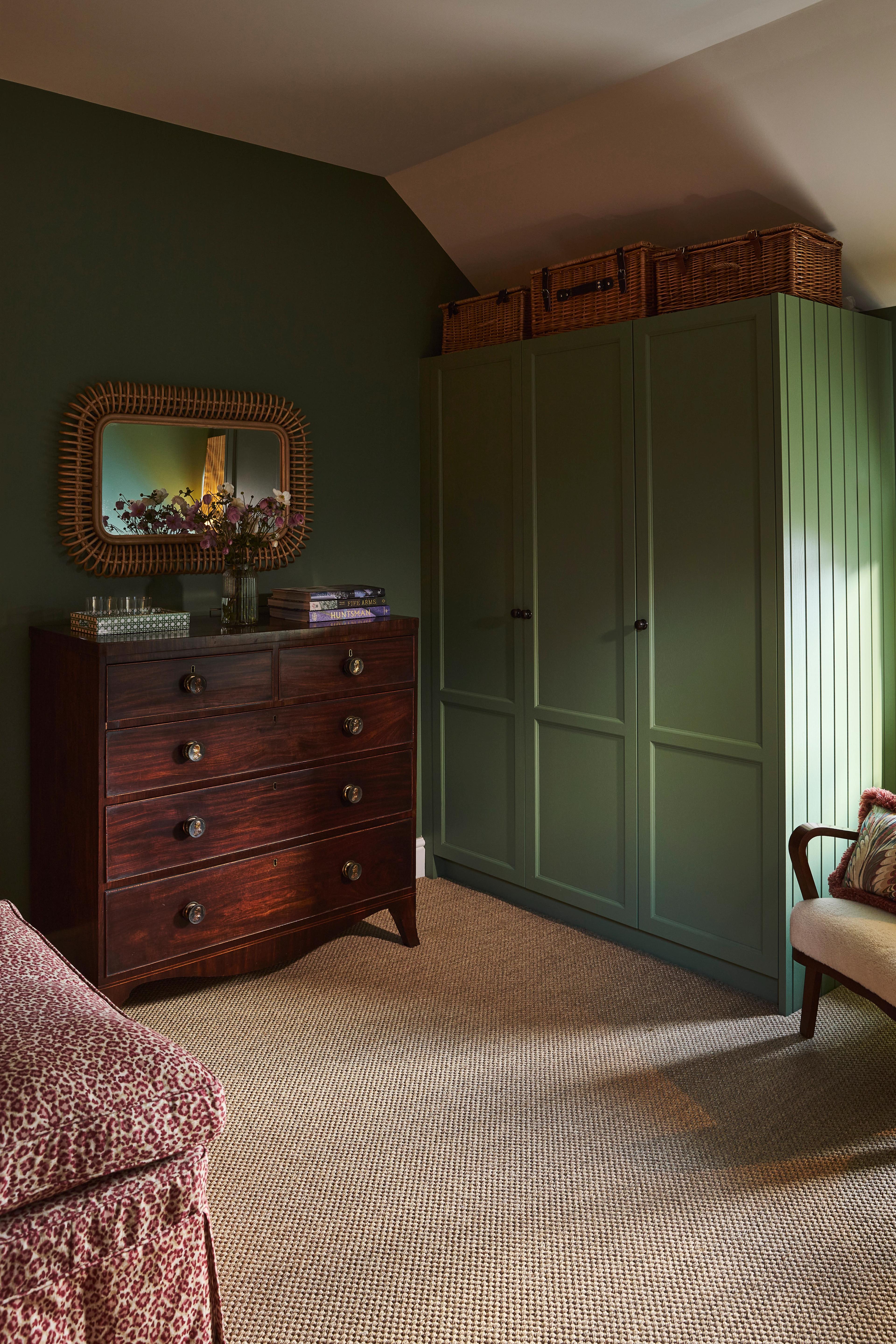 A cozy bedroom corner with olive green walls, a matching green wardrobe with baskets on top, a dark wood chest of drawers with a decorative mirror above, and a patterned armchair and carpet.