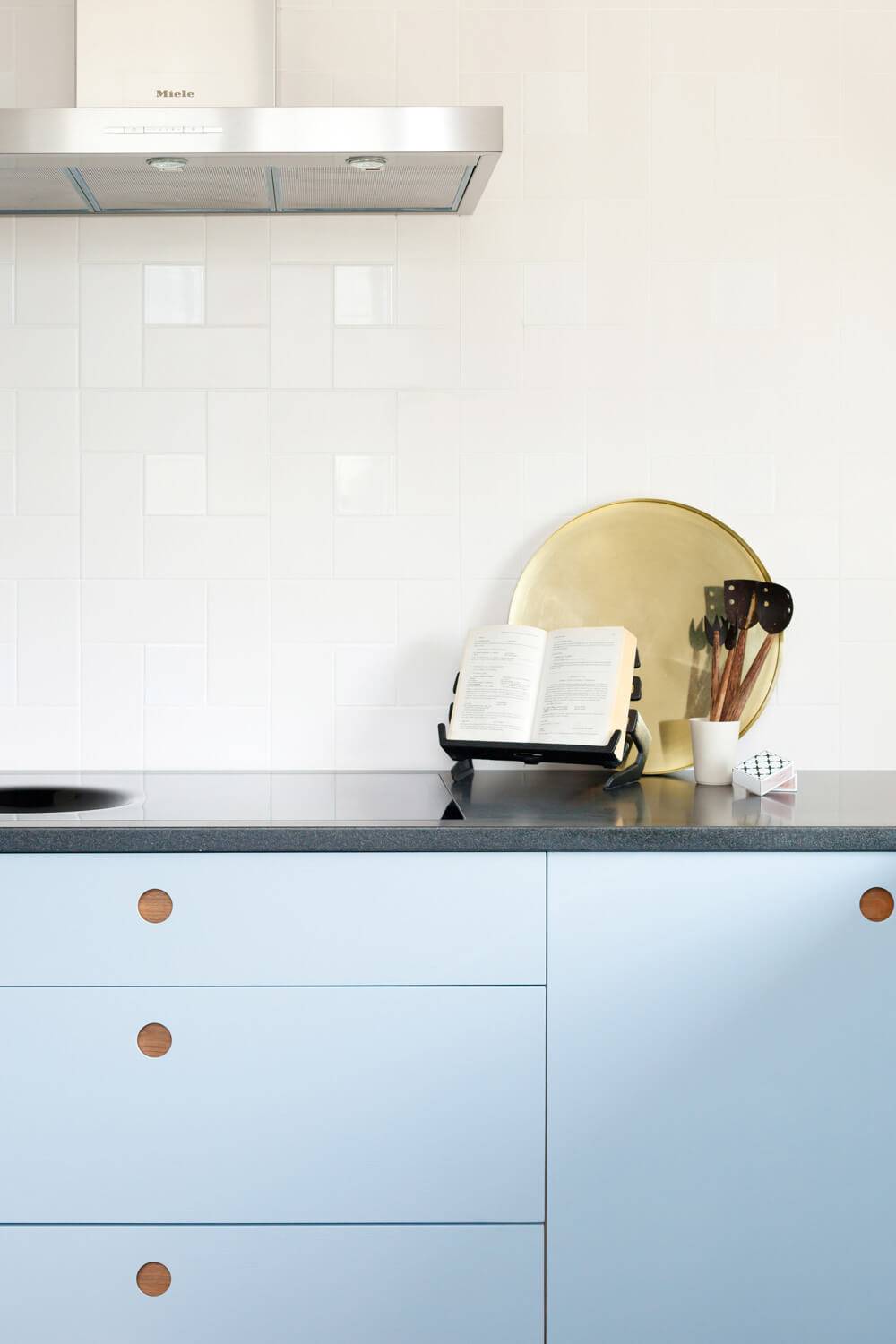 Modern kitchen with light blue cabinets featuring round wooden handles. A black countertop holds a plate rack with a cookbook, wooden utensils in a holder, and a white cup. A stainless steel range hood is mounted on white tile backsplash.