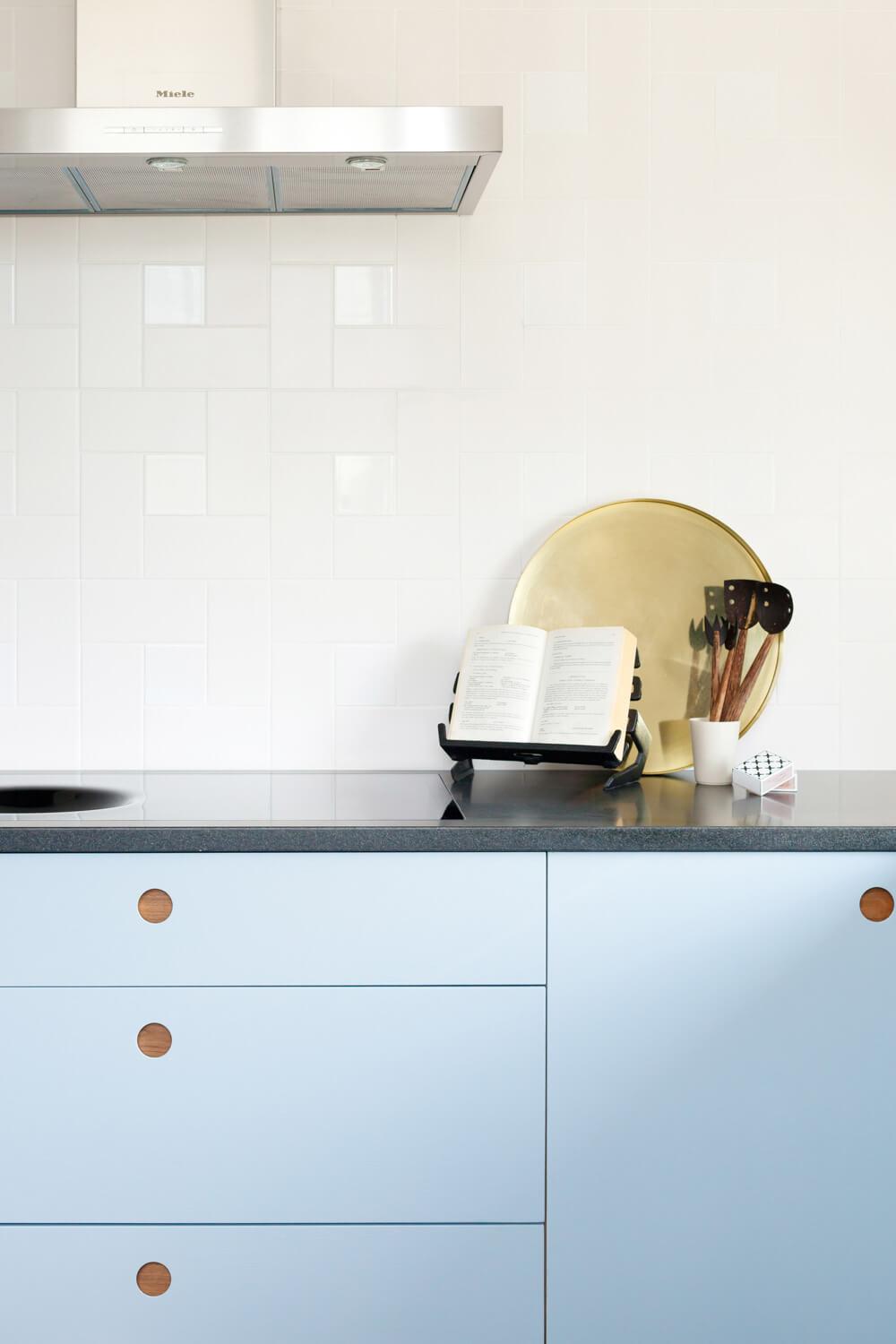 Modern kitchen with light blue cabinets featuring round wooden handles. A black countertop holds a plate rack with a cookbook, wooden utensils in a holder, and a white cup. A stainless steel range hood is mounted on white tile backsplash.