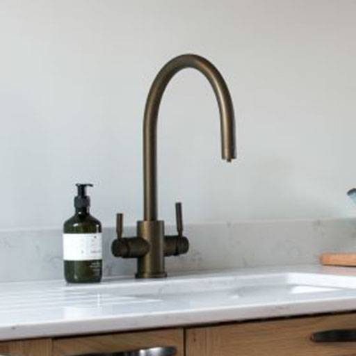 A modern kitchen sink with a curved bronze faucet, two handles, a white countertop, and a green soap dispenser on the left side.