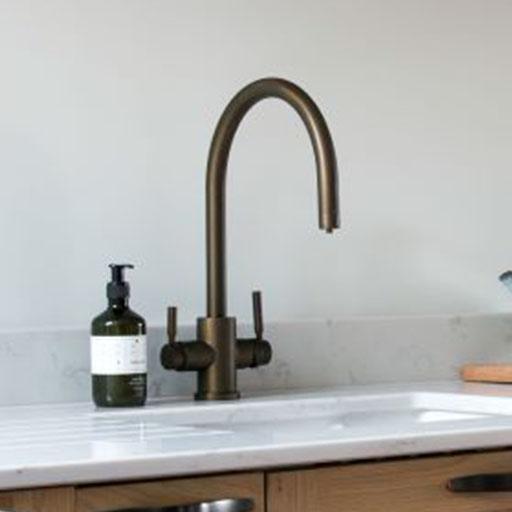 A modern kitchen sink with a curved bronze faucet, two handles, a white countertop, and a green soap dispenser on the left side.